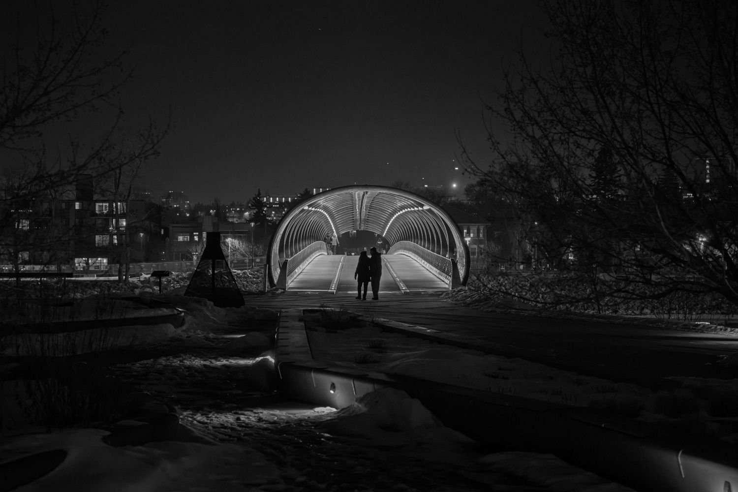Black and white night photo of couple standing under illuminated arched bridge in winter urban park.