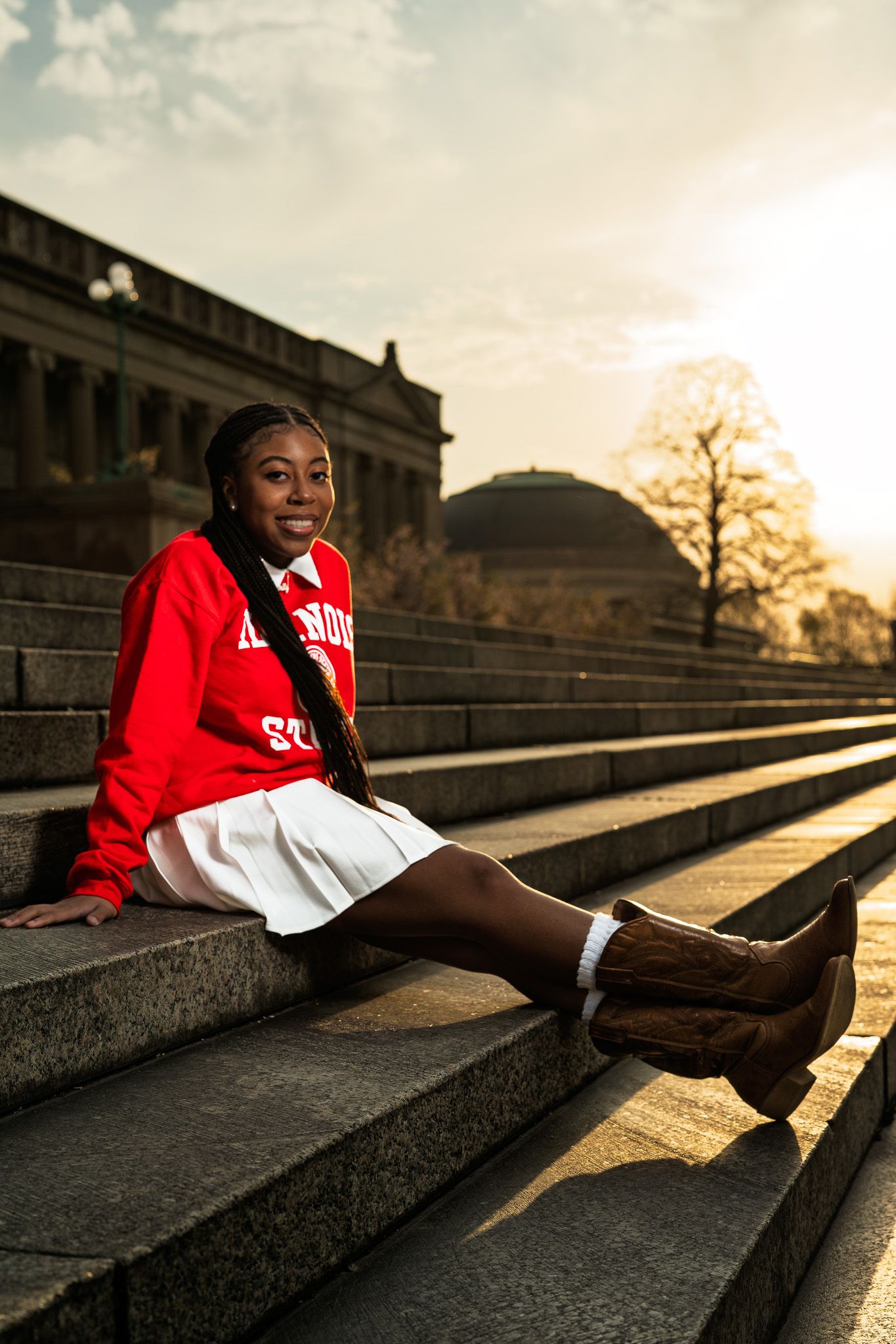 Young woman in red sweatshirt and white skirt sits smiling on stone steps with a domed building at golden hour.