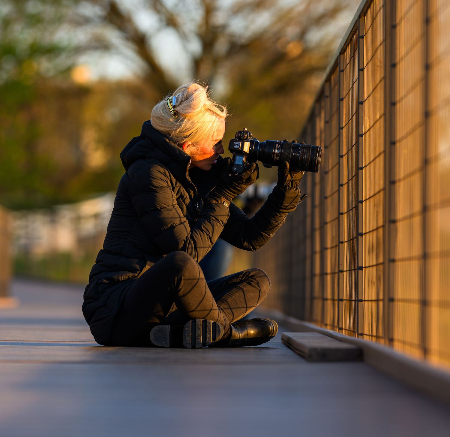 Photographer sitting on ground against fence, shooting with DSLR camera at golden hour in winter coat and beanie.