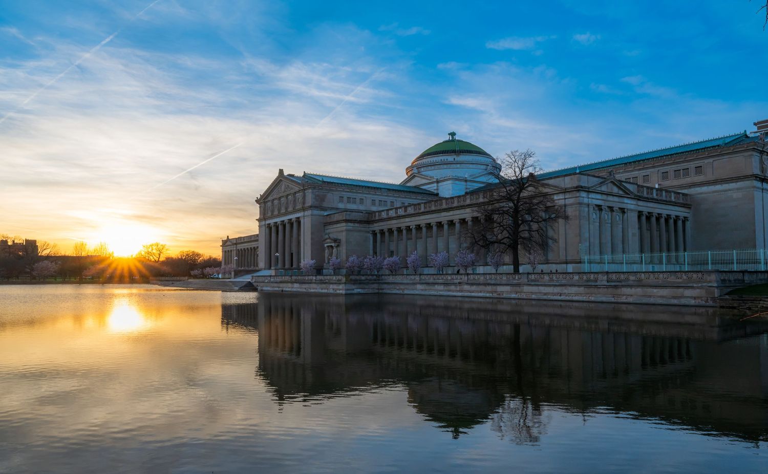 Sunset reflecting on a lagoon beside a grand neoclassical museum building with golden light and blue sky.