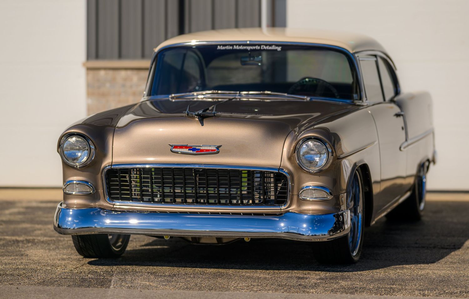 A custom 1955 Chevy Bel Air in bronze and blue two-tone paint sits outside a building.