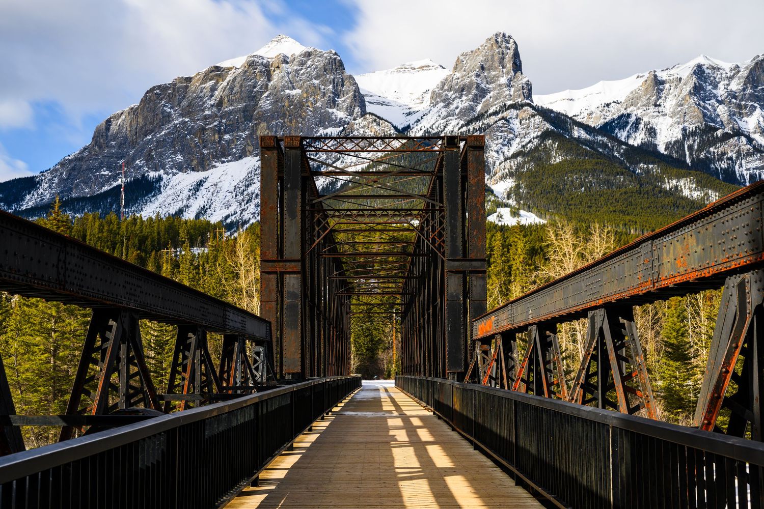 Rustic iron railway bridge with a vanishing point perspective framing snow-capped Rocky Mountains and evergreen forest.