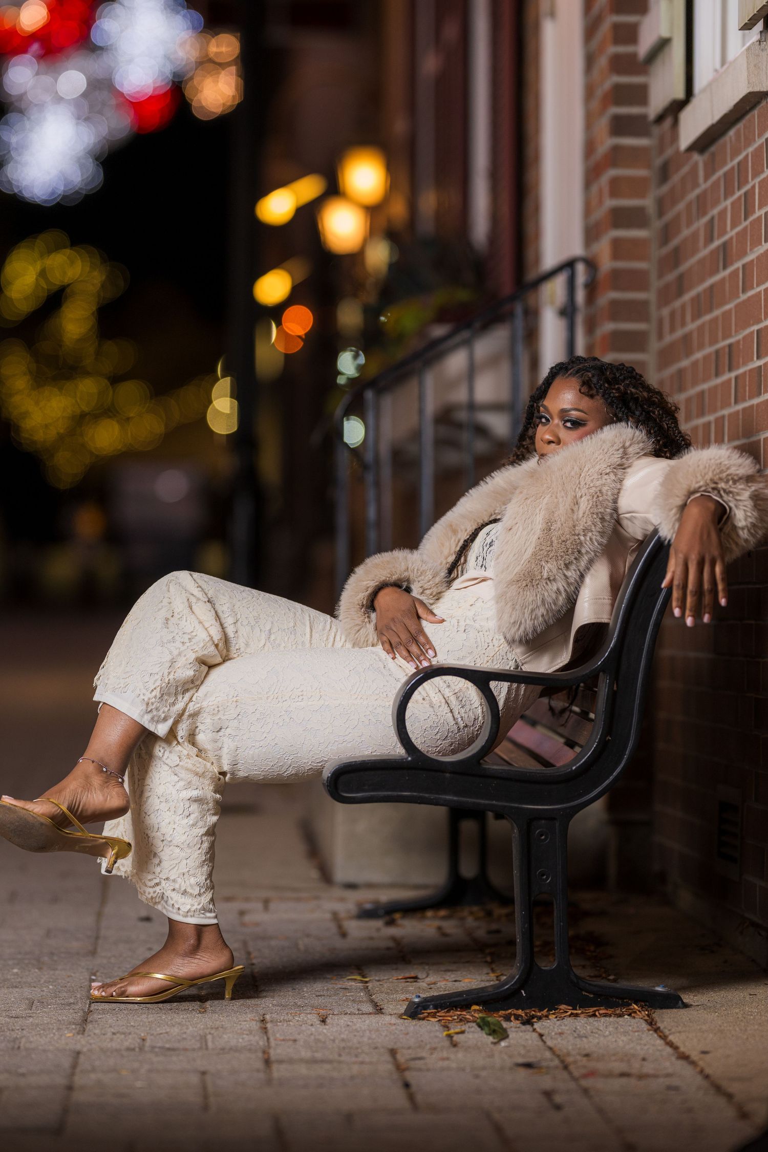 A stylish woman in white lace outfit and fur coat poses on a black bench at night on a city sidewalk.