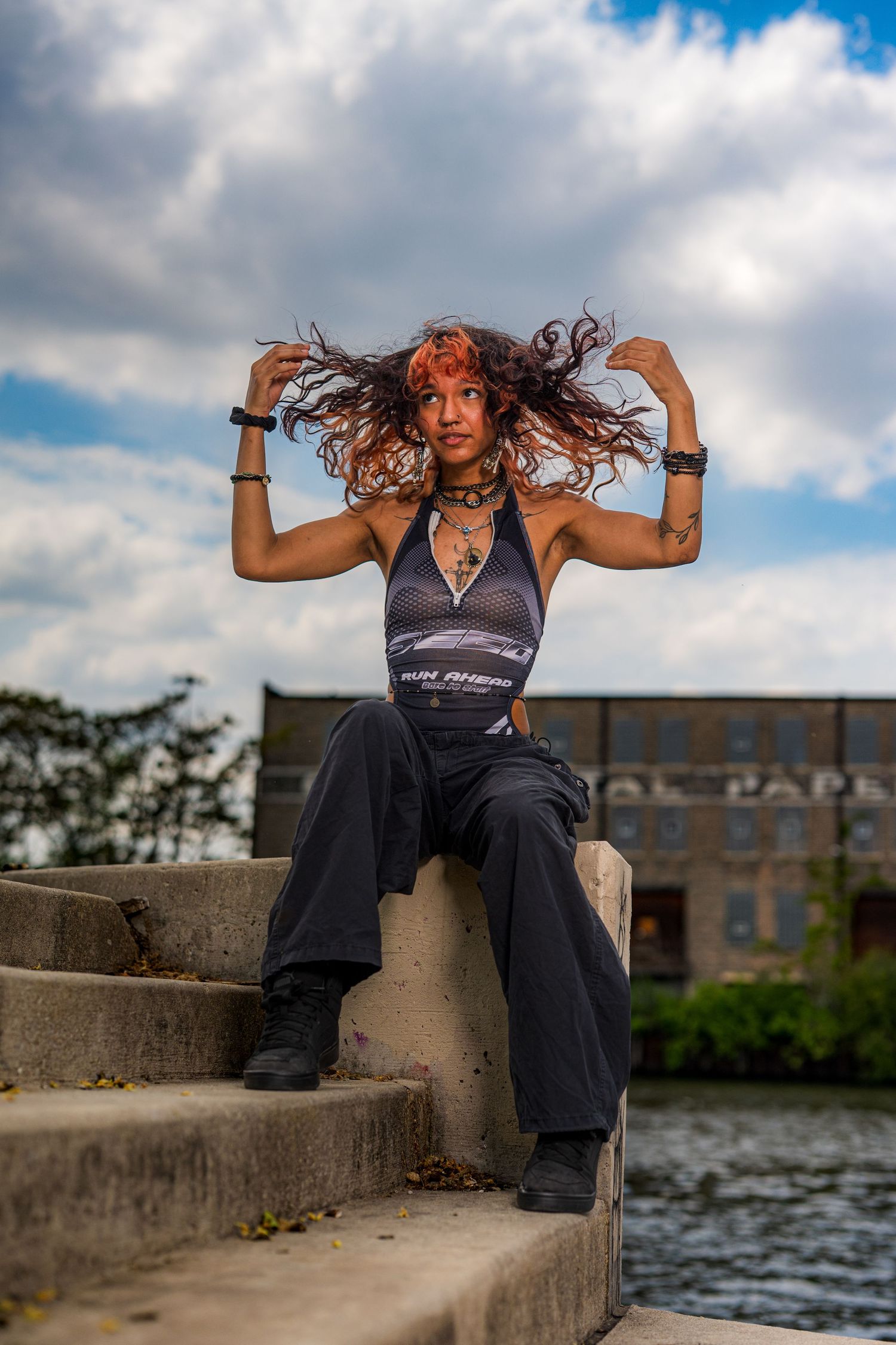 Woman with red hair poses on concrete steps outdoors, wearing a halter top and black pants, with a cloudy sky behind her.