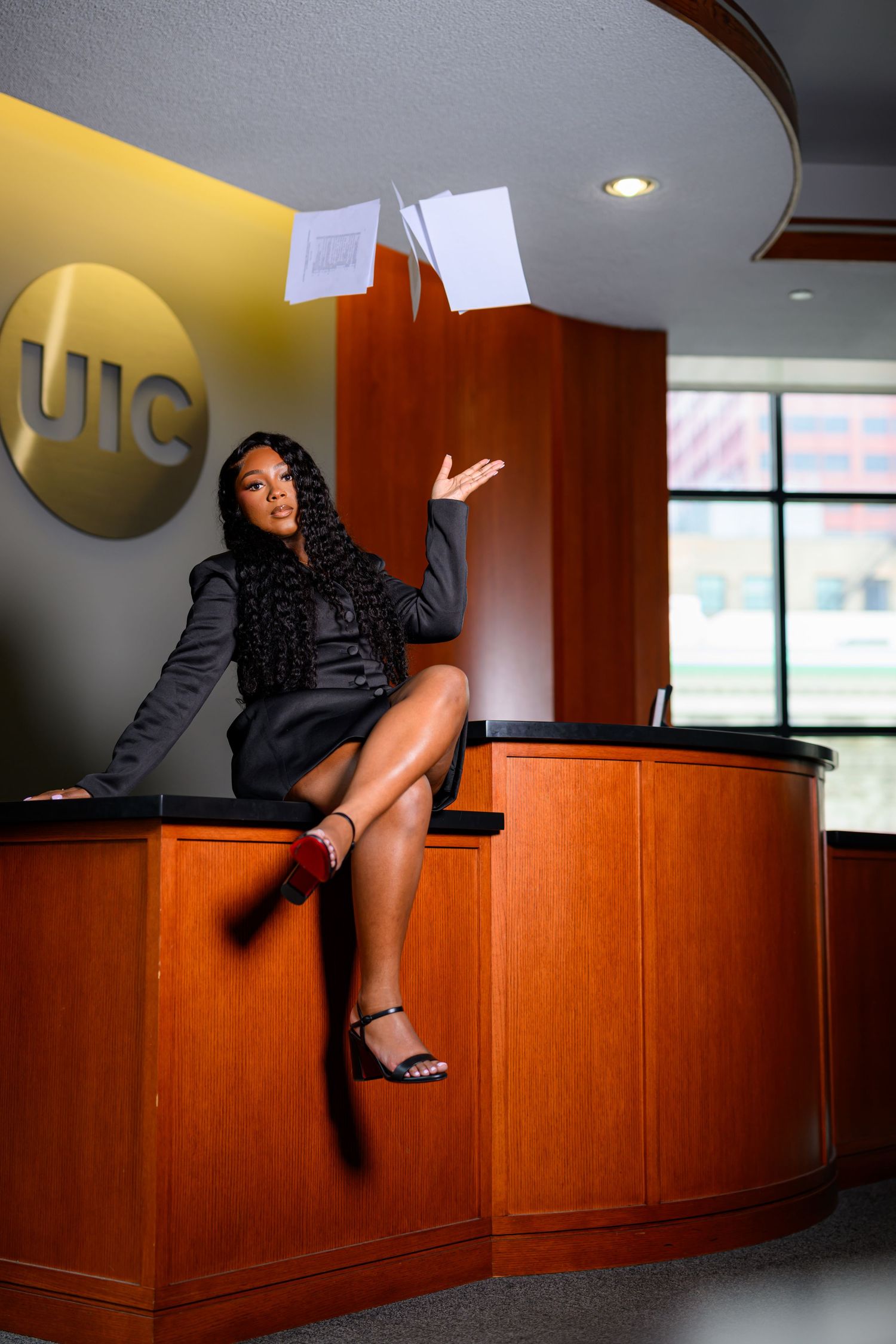 A young woman in a black outfit sits on a UIC reception desk, tossing papers in the air with a confident smile.