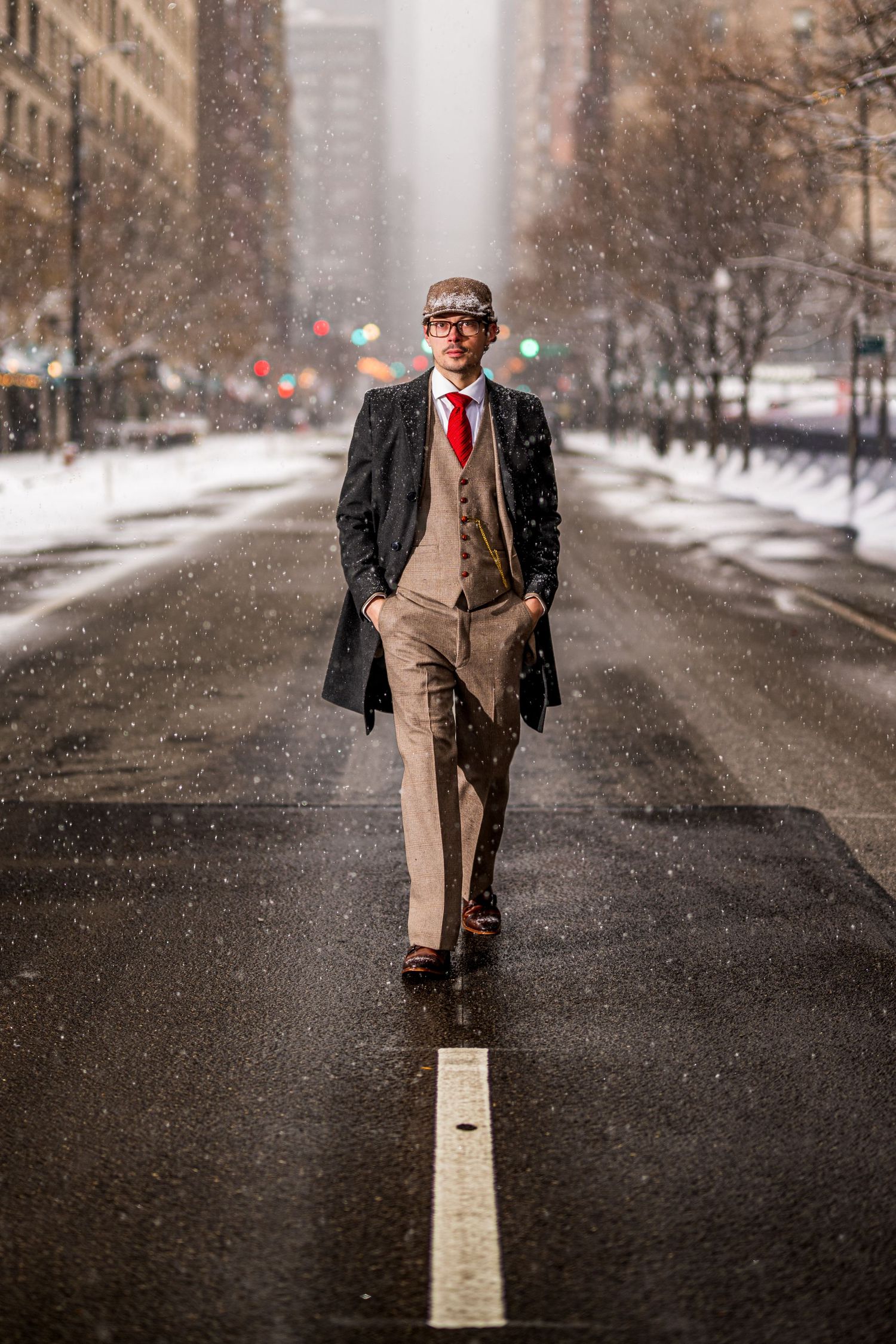 A stylish man in a tweed suit, red tie, and long coat walks confidently down a snowy urban street.