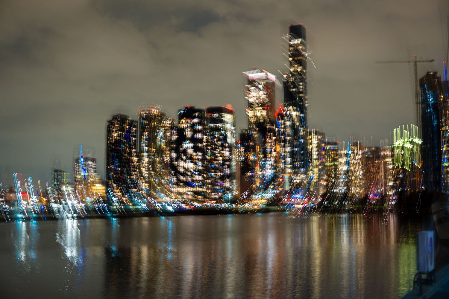 Blurred city skyline reflecting colorful lights on water at night, creating an abstract bokeh effect.