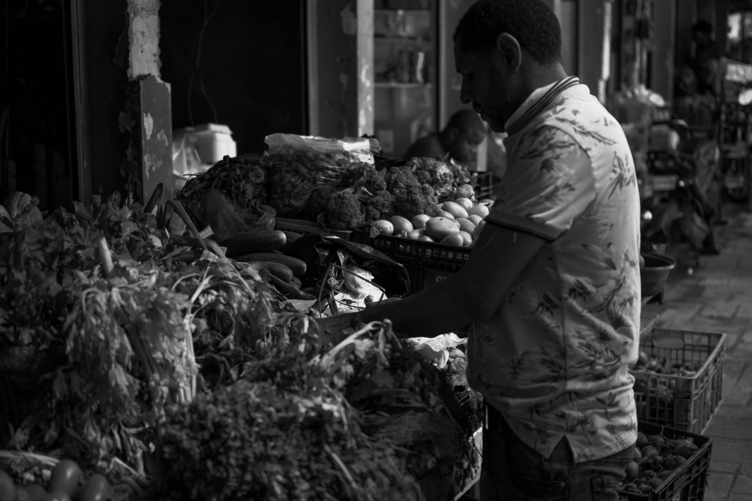 Black and white photo of a man browsing fresh vegetables at an outdoor market stall.