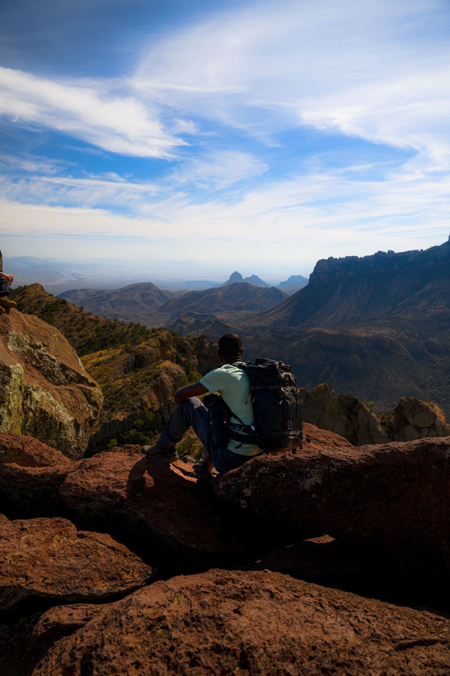 Hiker with backpack sitting on rocky summit overlooking vast mountain valley landscape under blue sky in Big Bend National Park.