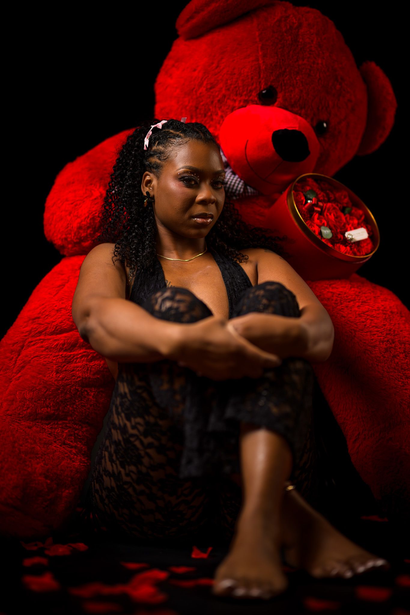 Woman in black lace dress sits against large red teddy bear with rose petals and candy on dark background.