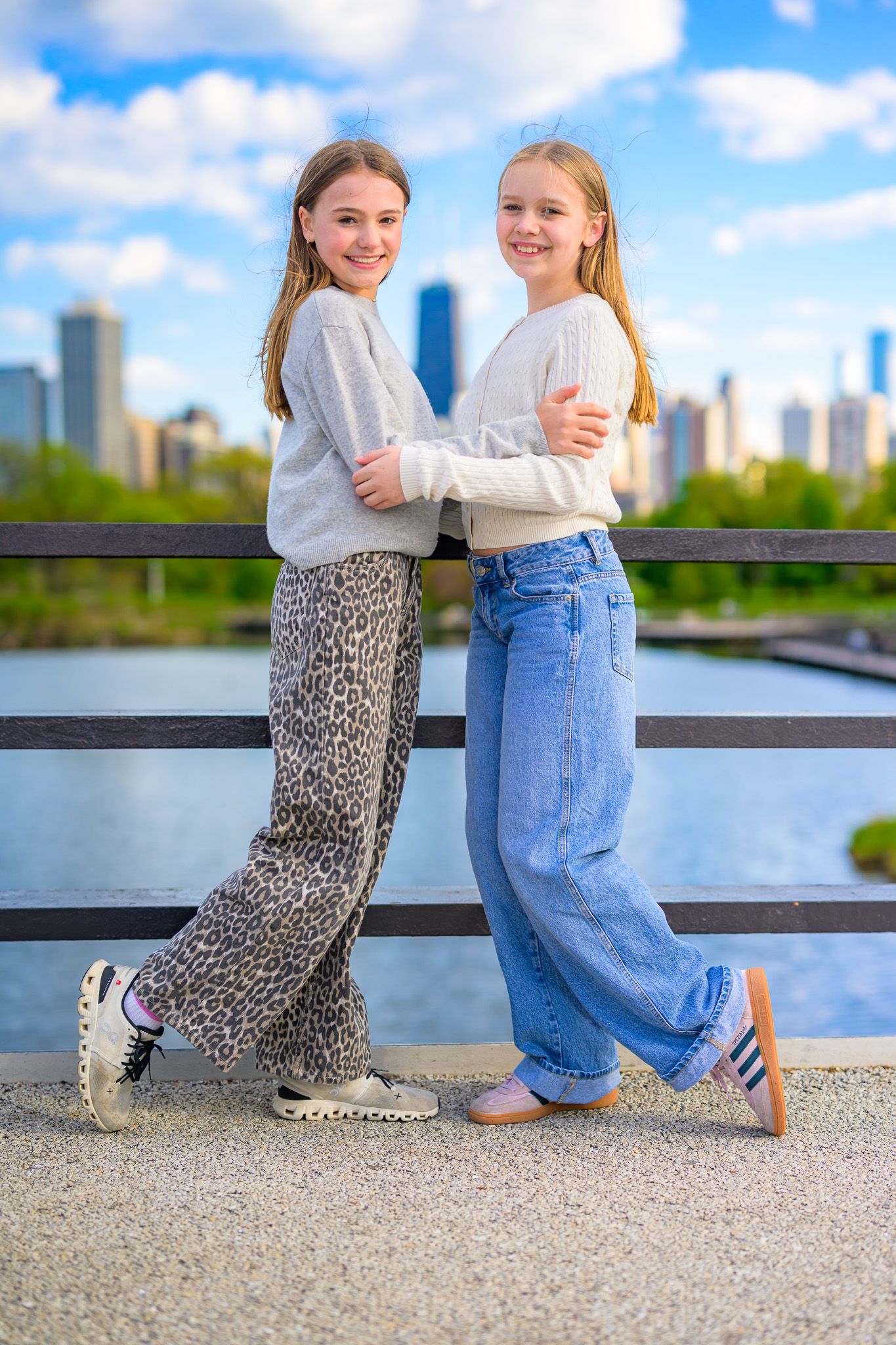 Two smiling girls in wide-leg pants pose together on a bridge with a city skyline backdrop.