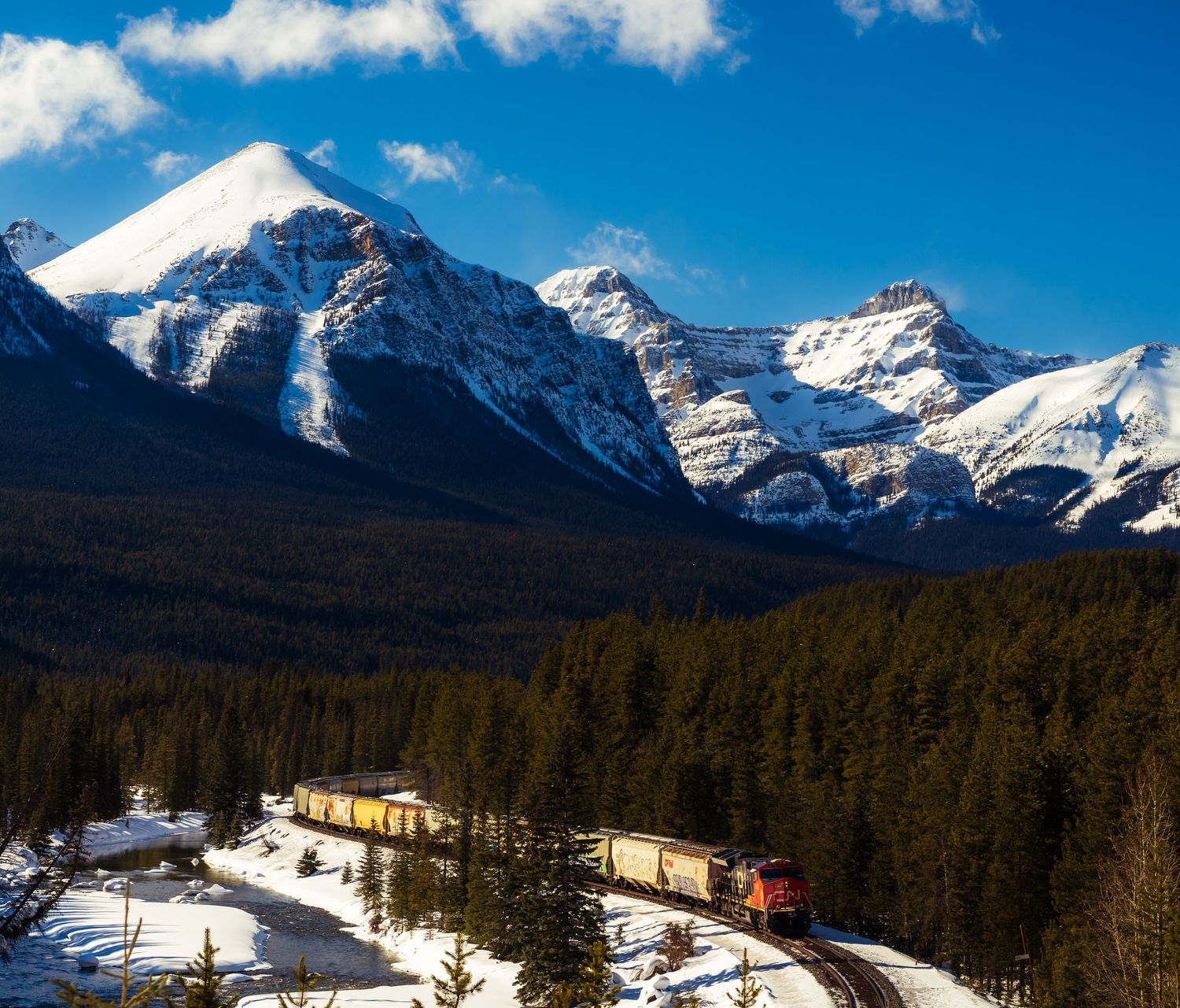 A red freight train winds through a snowy Canadian Rockies forest with majestic snow-capped mountain peaks under a blue sky.