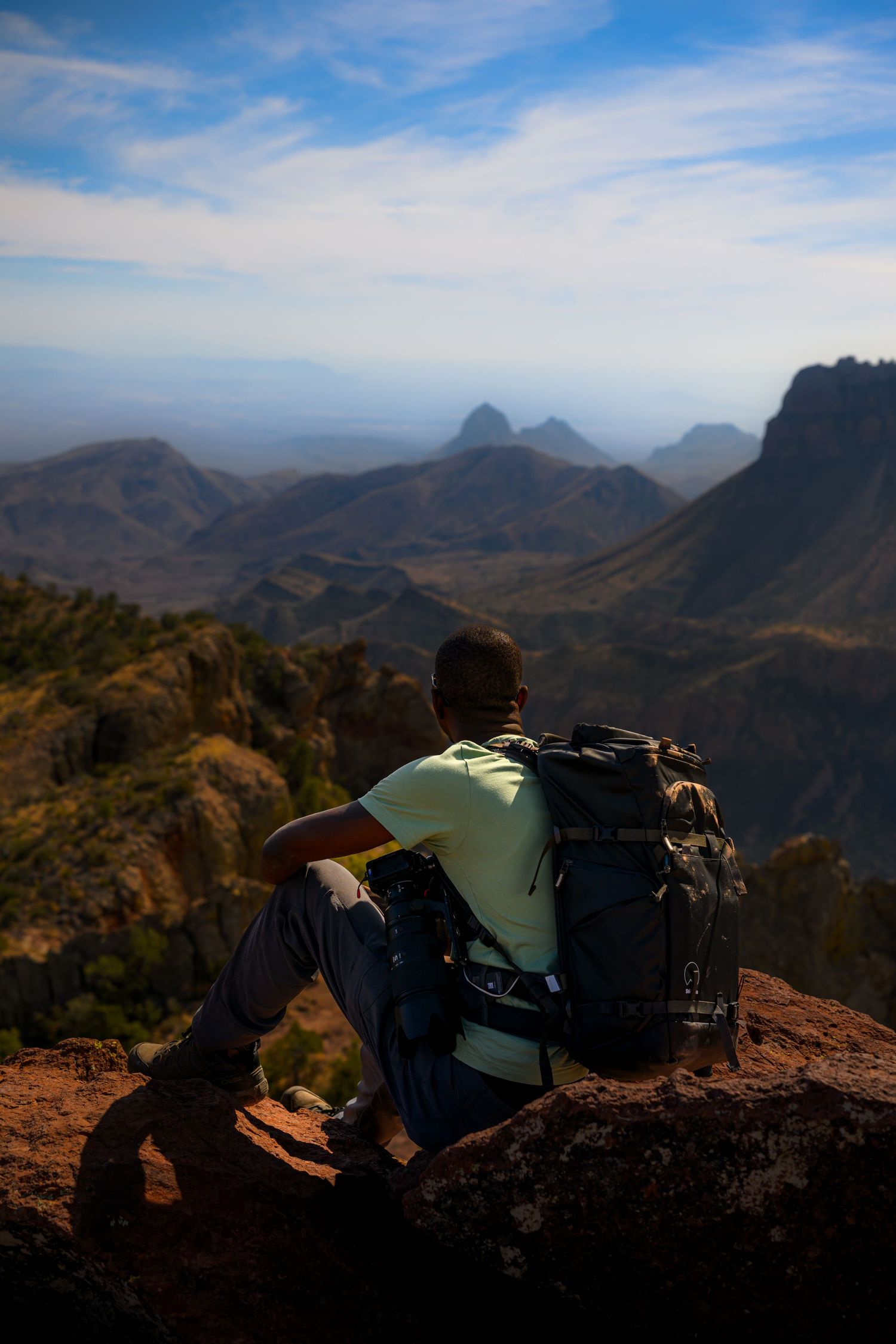 Hiker with backpack sitting on rocky summit overlooking vast mountain landscape under blue sky.