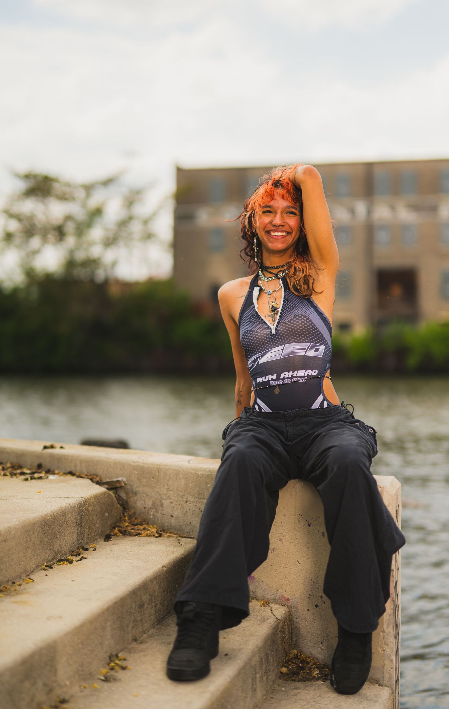 Smiling woman with red hair sits on concrete steps by a river, wearing a graphic halter top and black wide-leg pants.