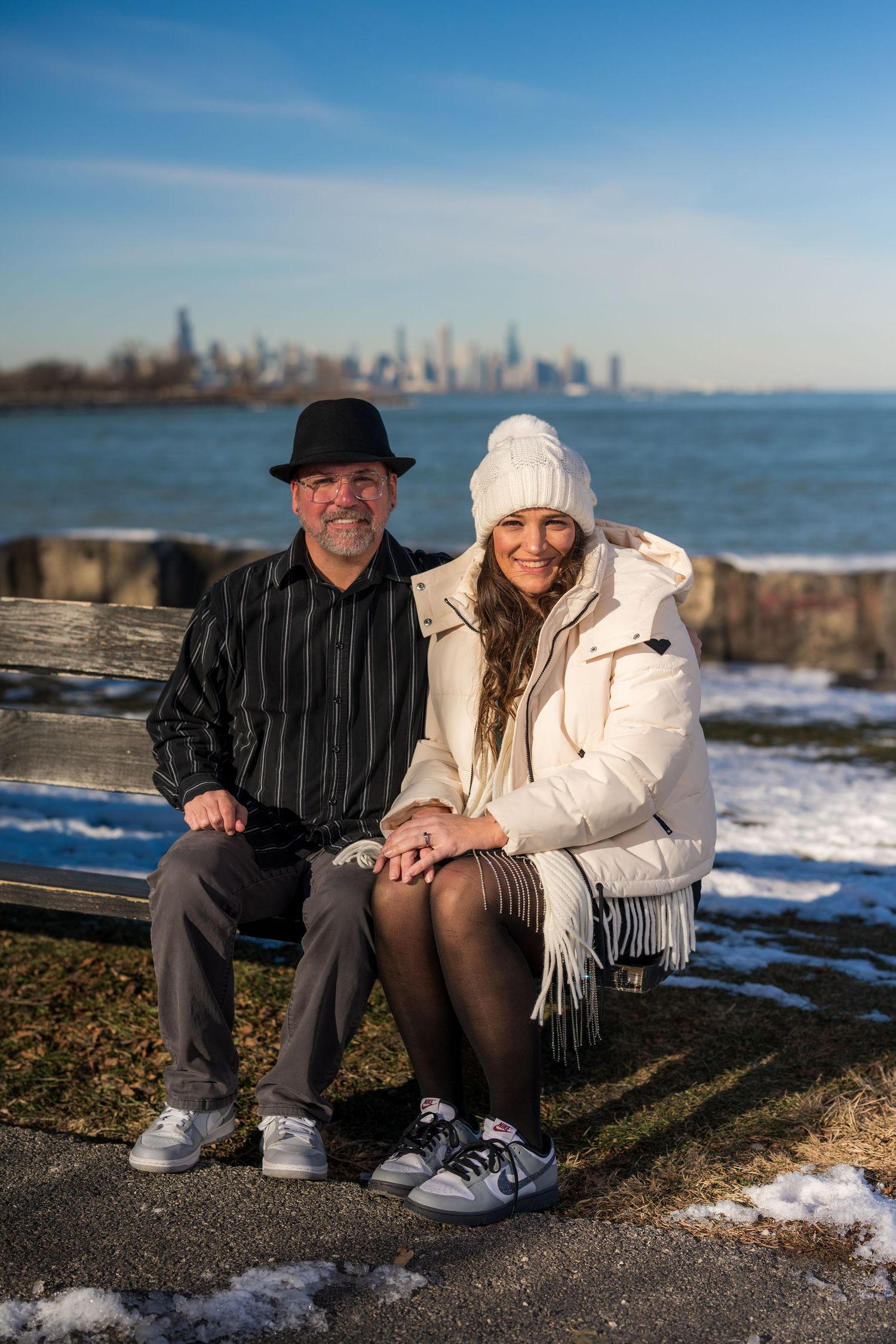 Couple sitting on bench by lakeside with city skyline in winter, wearing hats and white puffer jacket.