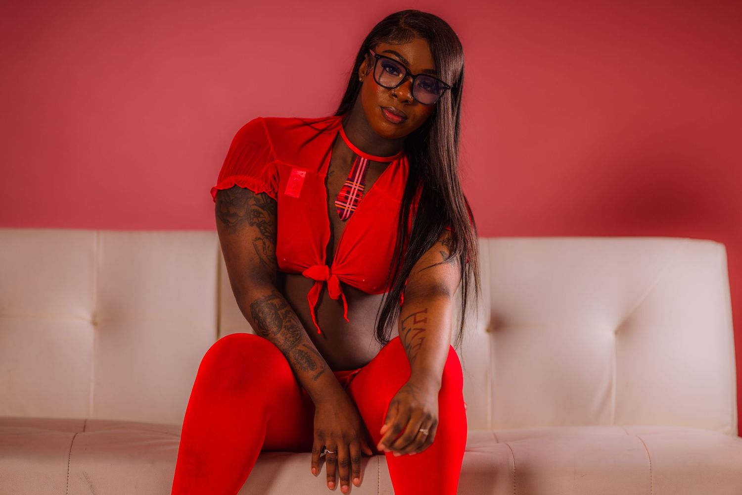 A woman in a red outfit and glasses sits confidently on a white couch against a pink background.