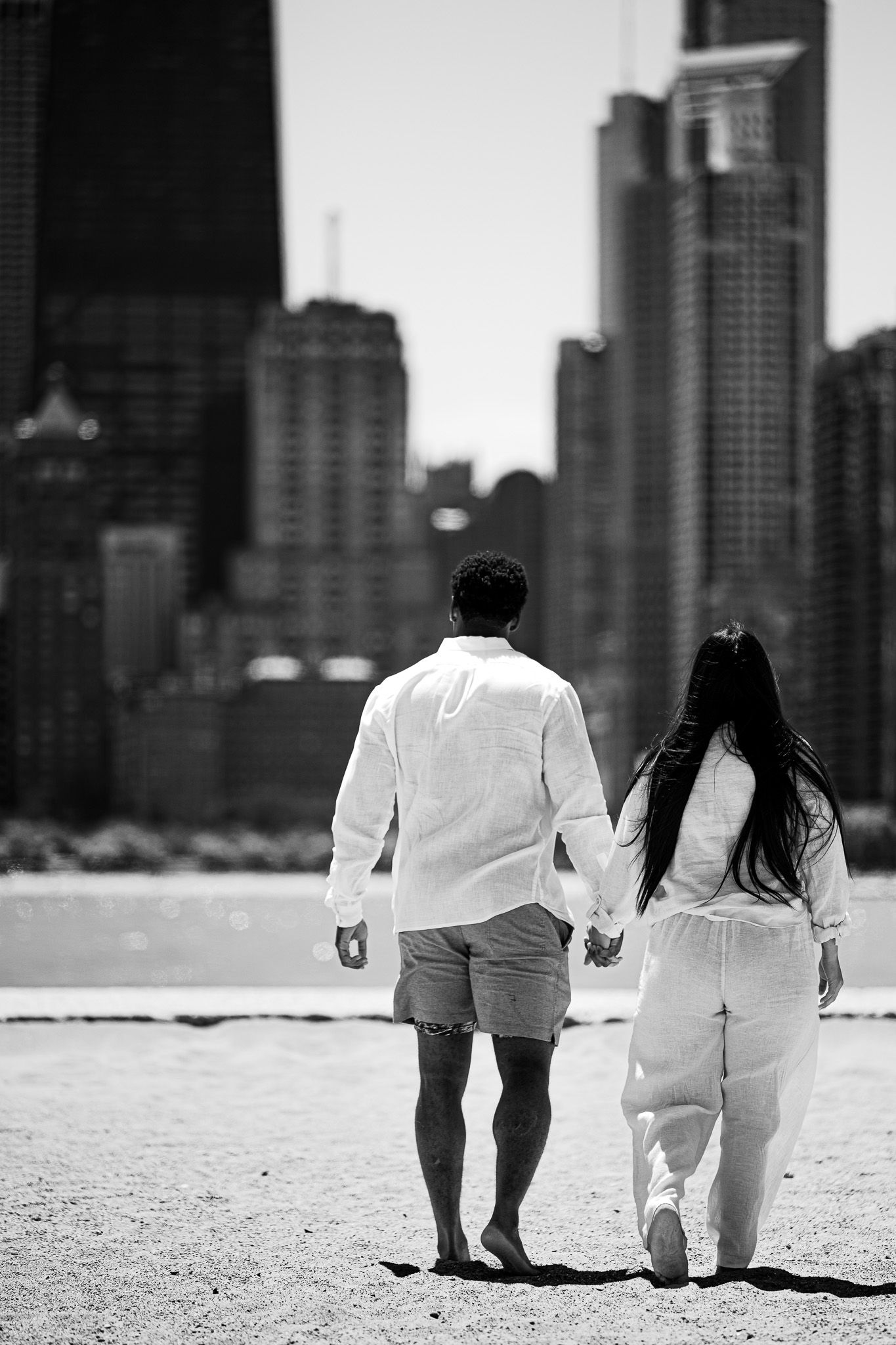 Black and white photo of couple holding hands walking toward city skyline on beach.