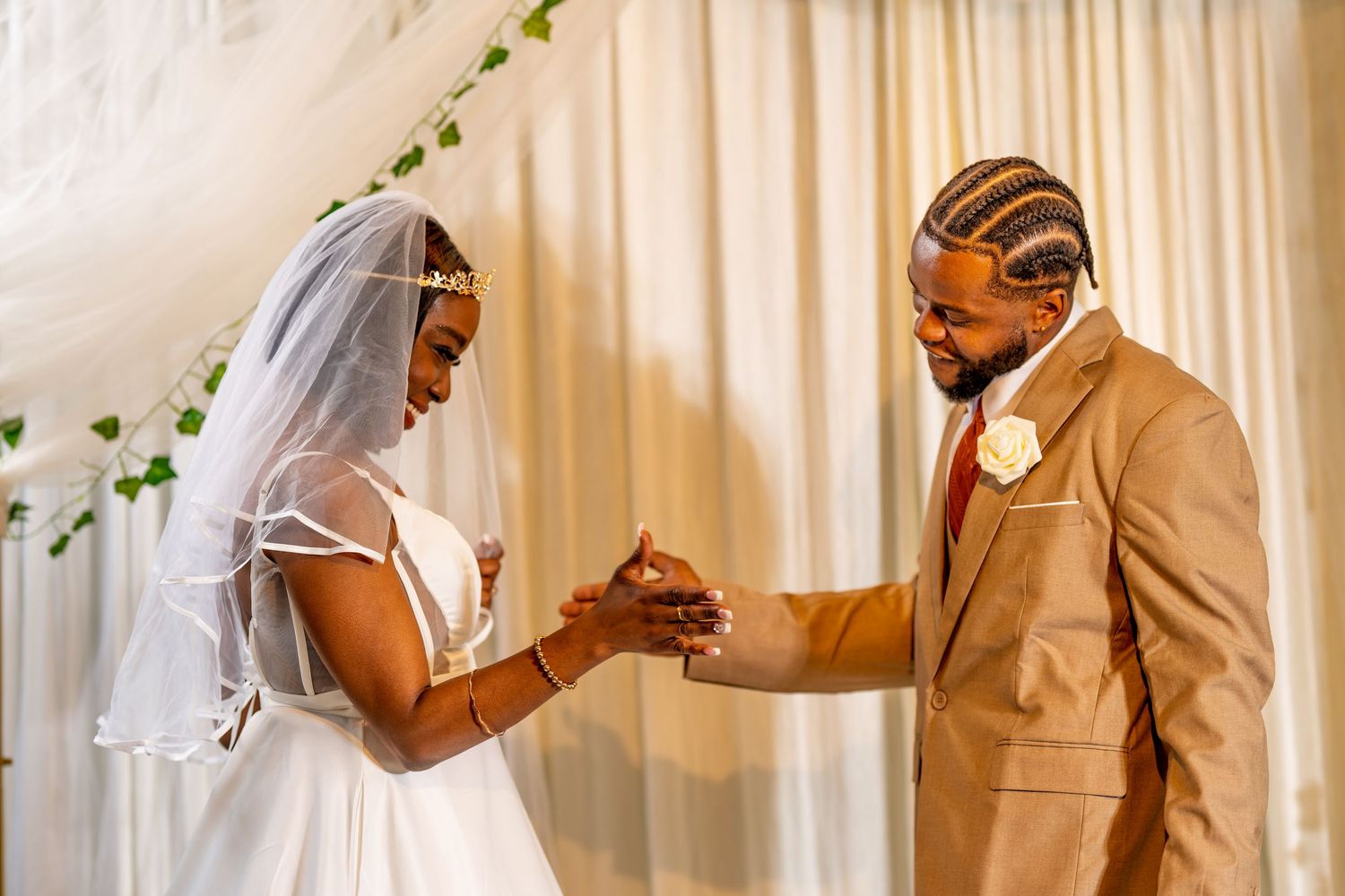 Bride and groom exchange rings at wedding ceremony, bride in white gown and veil, groom in tan suit.