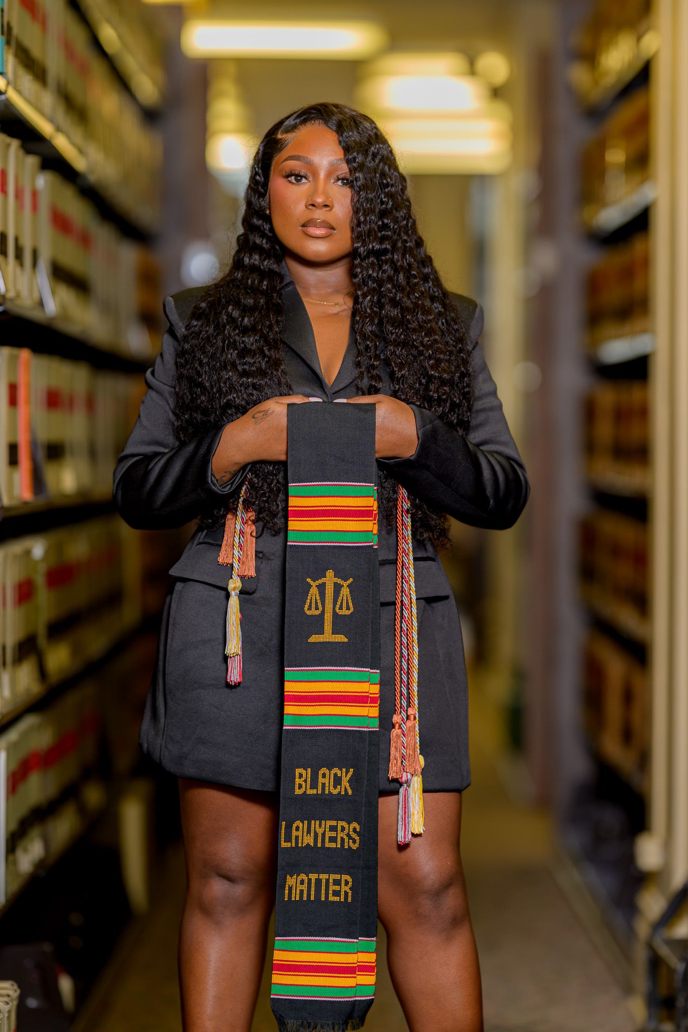 A Black law school graduate holds a 'Black Lawyers Matter' kente stole with scales of justice in a library.