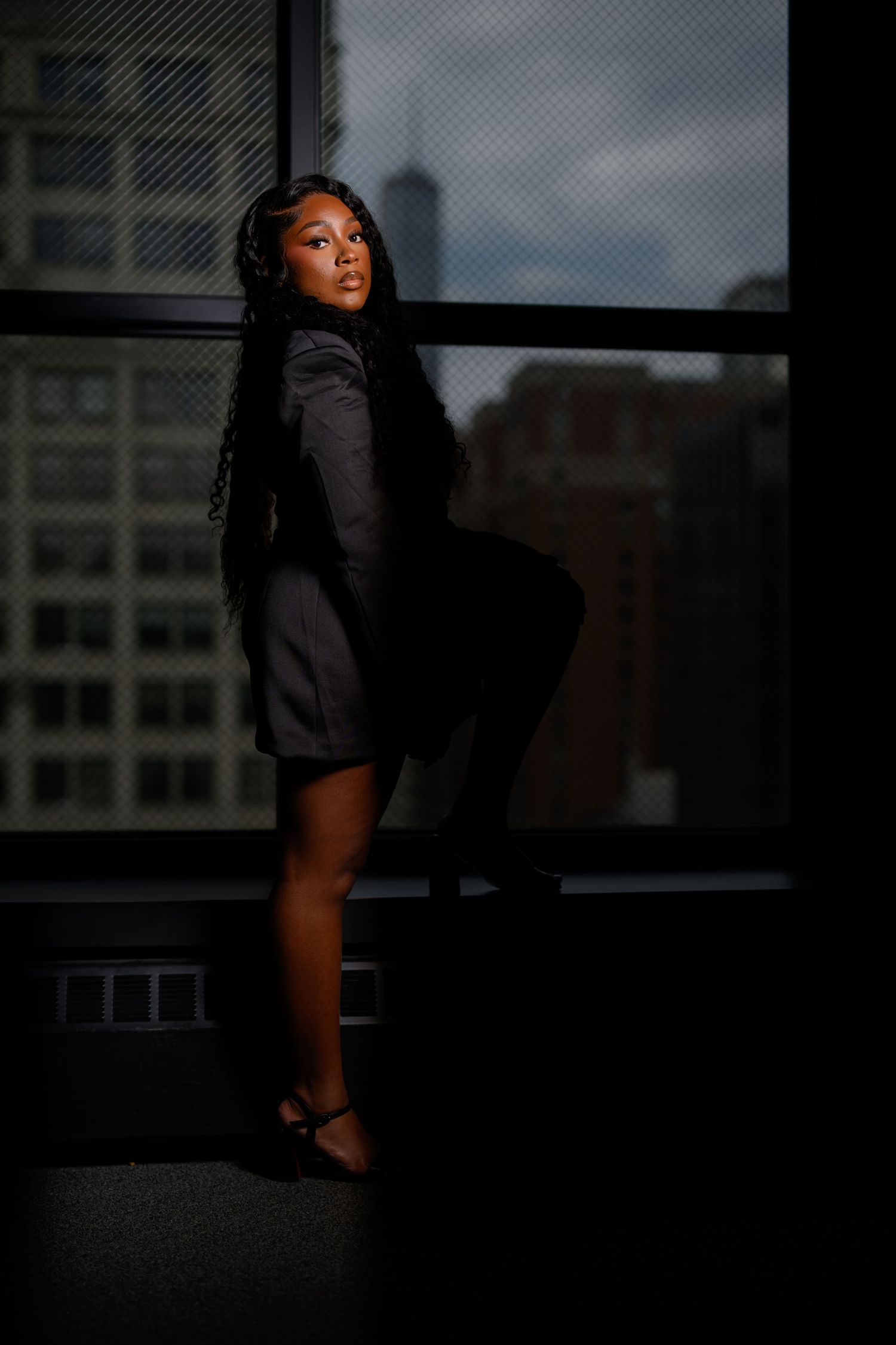 A young Black woman in a gray blazer dress poses confidently by large windows with a city skyline backdrop.
