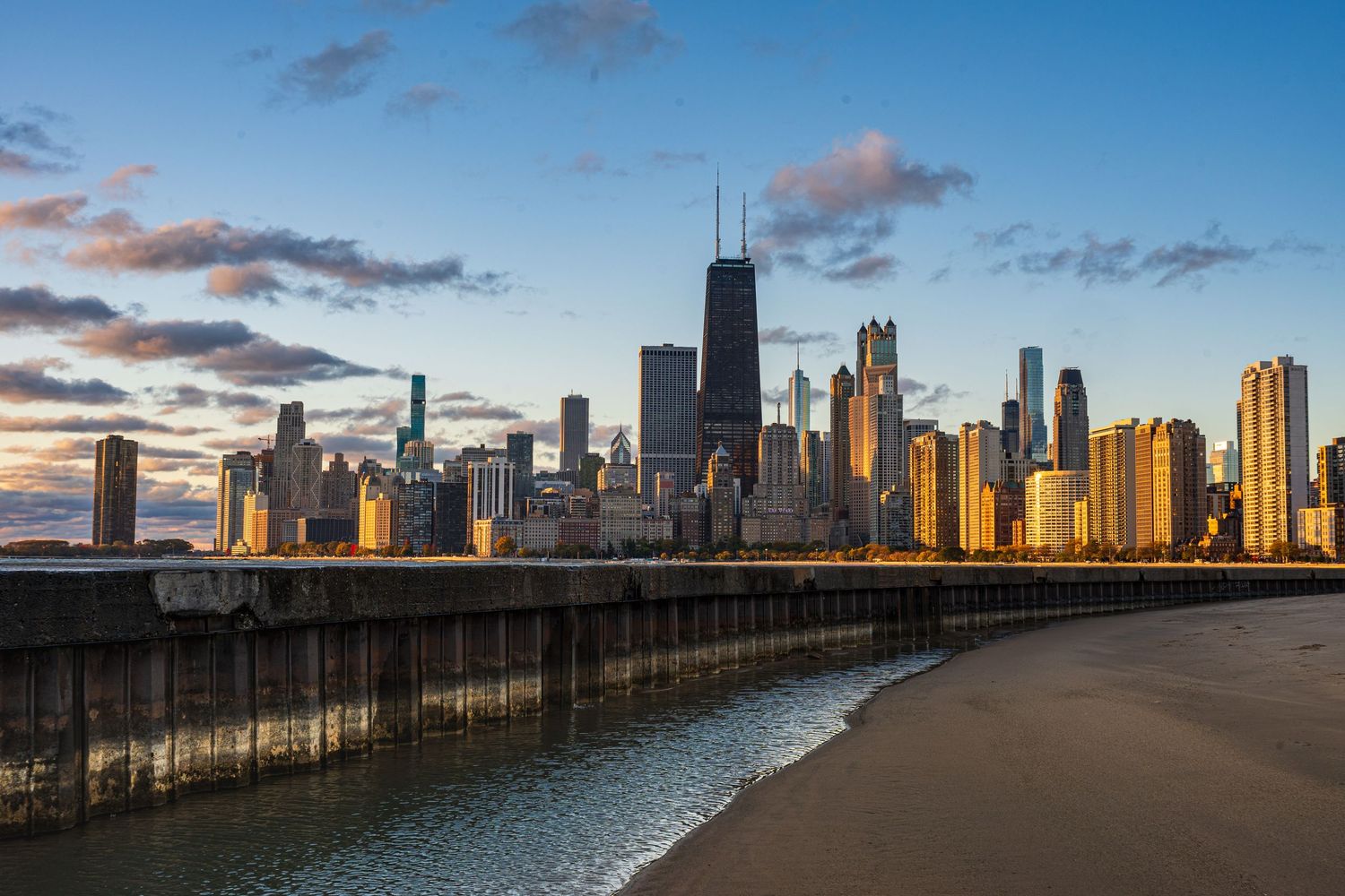 Chicago skyline at sunset viewed from a sandy beach with a concrete pier, featuring the iconic John Hancock Center.