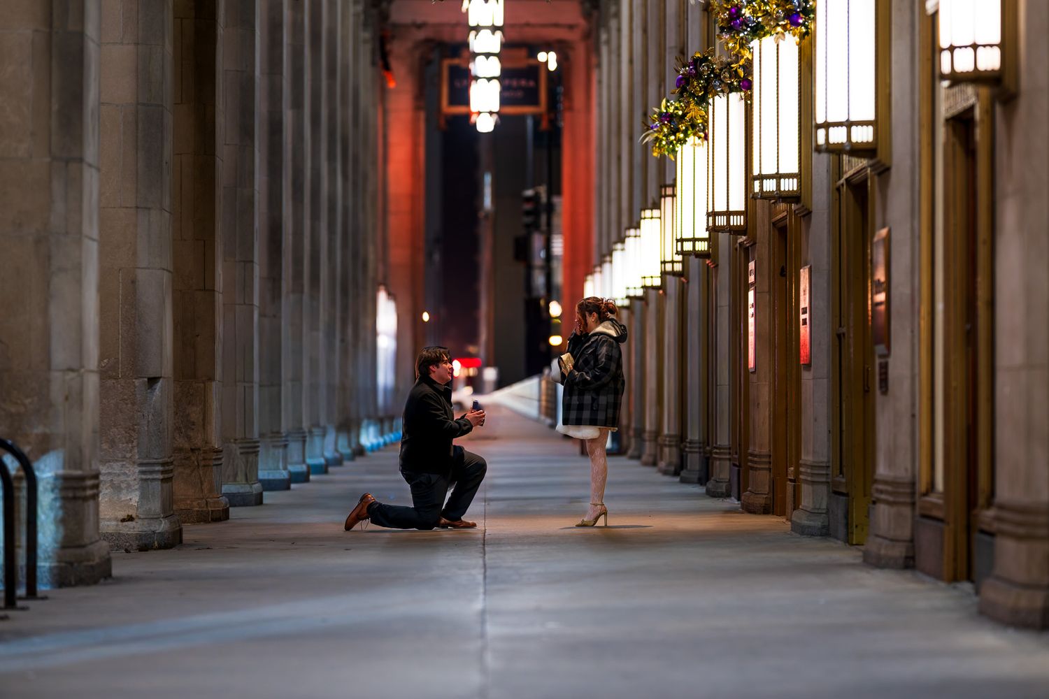 Man on one knee proposing to woman in elegant illuminated arcade colonnade at night.