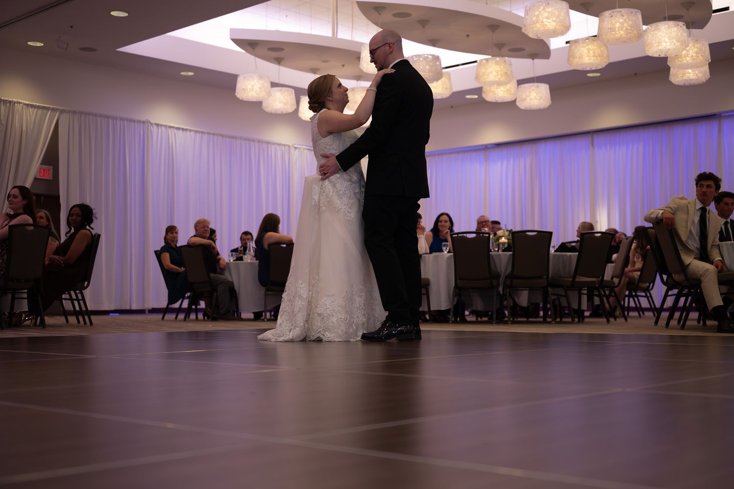 Bride and groom share their first dance at elegant wedding reception with soft lighting and guests seated in background.
