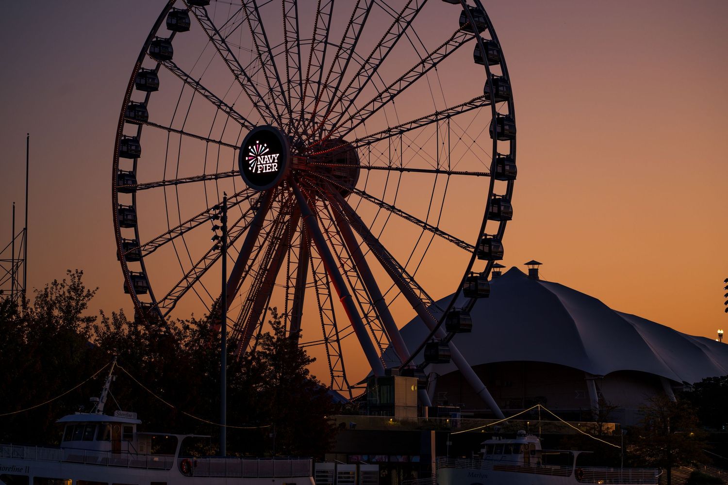 Silhouette of a large Ferris wheel against a vibrant orange and purple sunset sky at a waterfront amusement park.