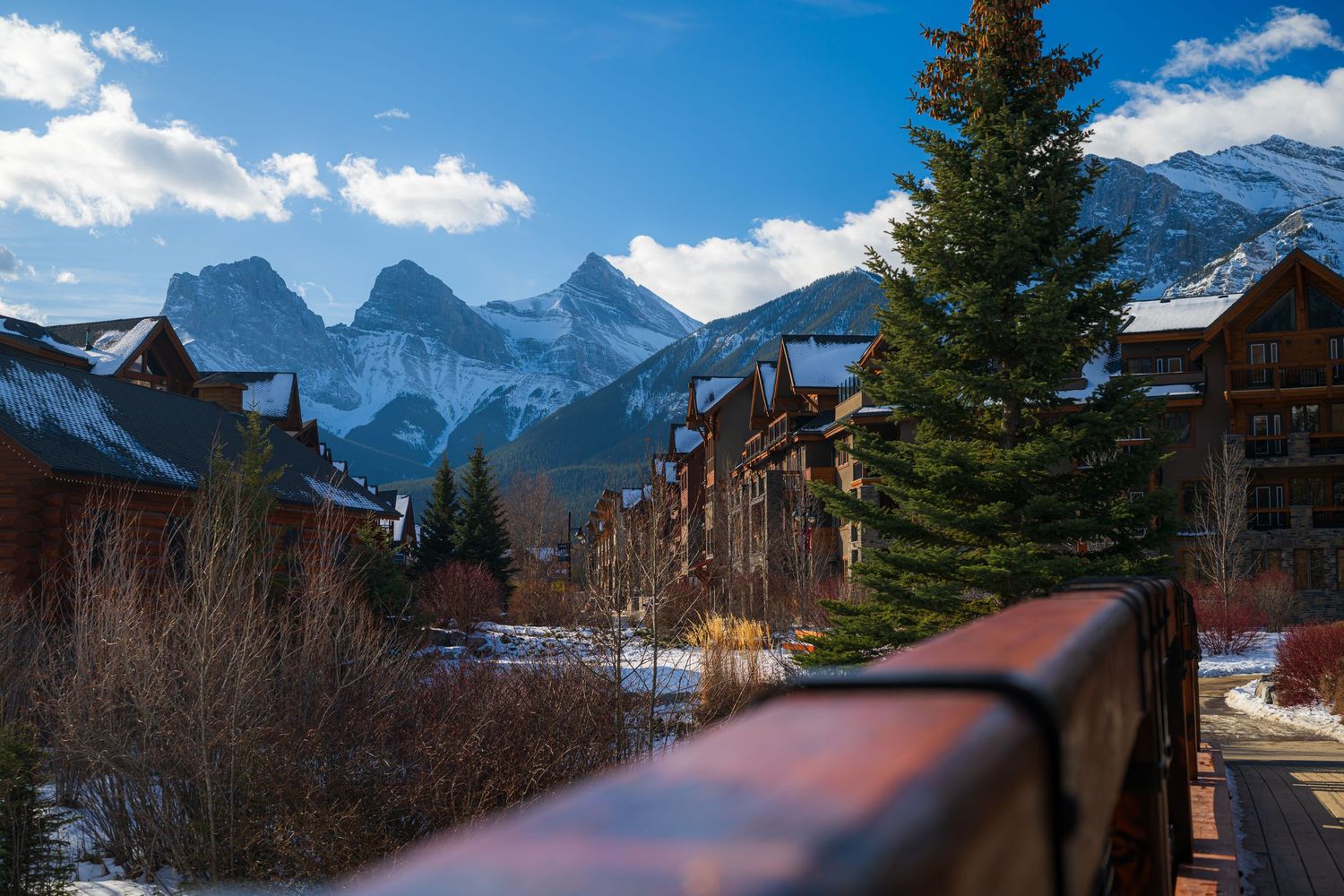 Snow-dusted mountain peaks rise behind rustic lodge buildings and evergreen trees in a scenic winter resort town.