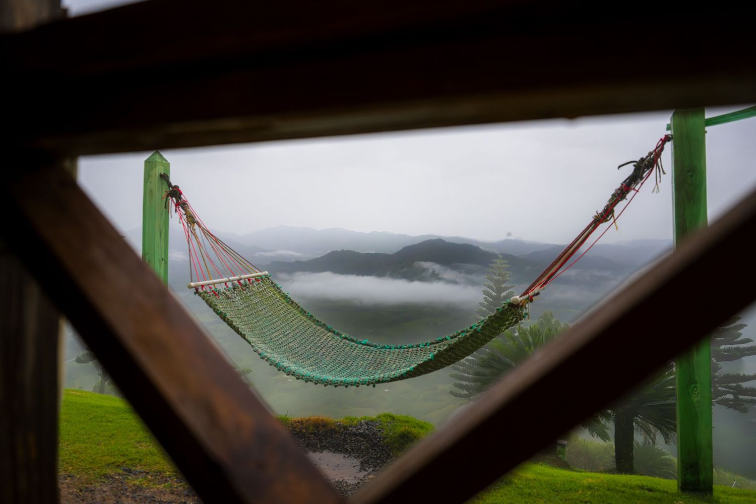 A rope hammock strung between green posts overlooks misty mountain valleys through a rustic wooden window frame.