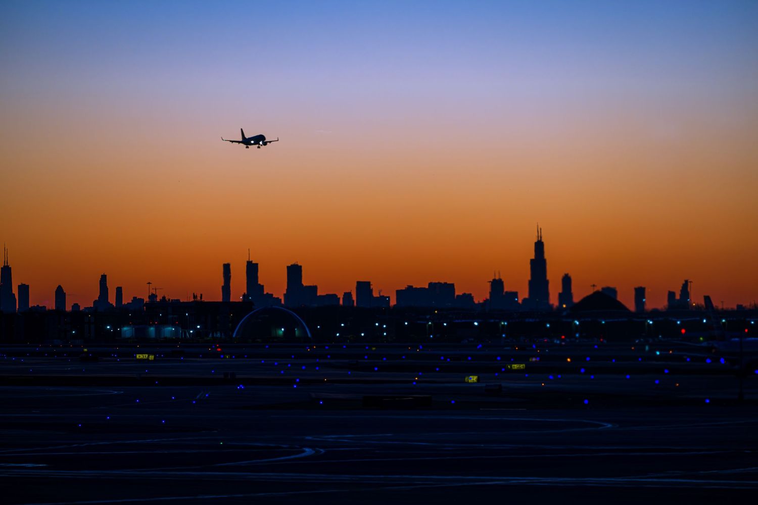 Airplane silhouette flying over Chicago skyline at sunset with vibrant orange and blue gradient sky.