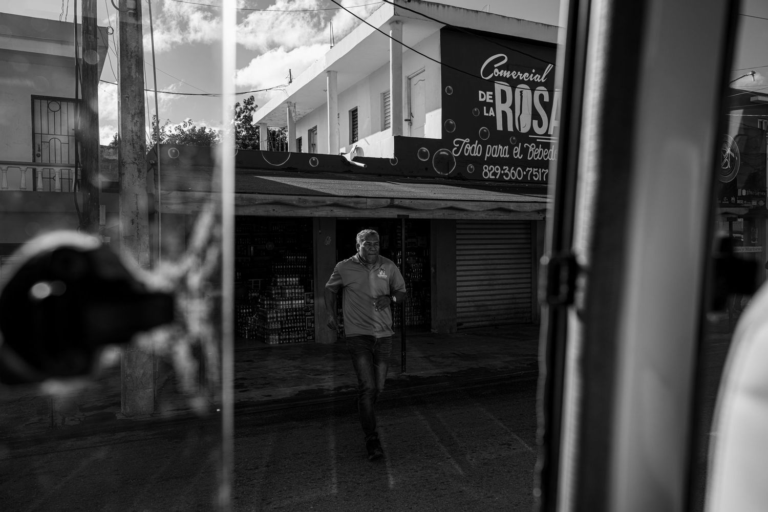 Black and white street photo of a man jogging past storefronts, captured through a reflective glass window in an urban setting.