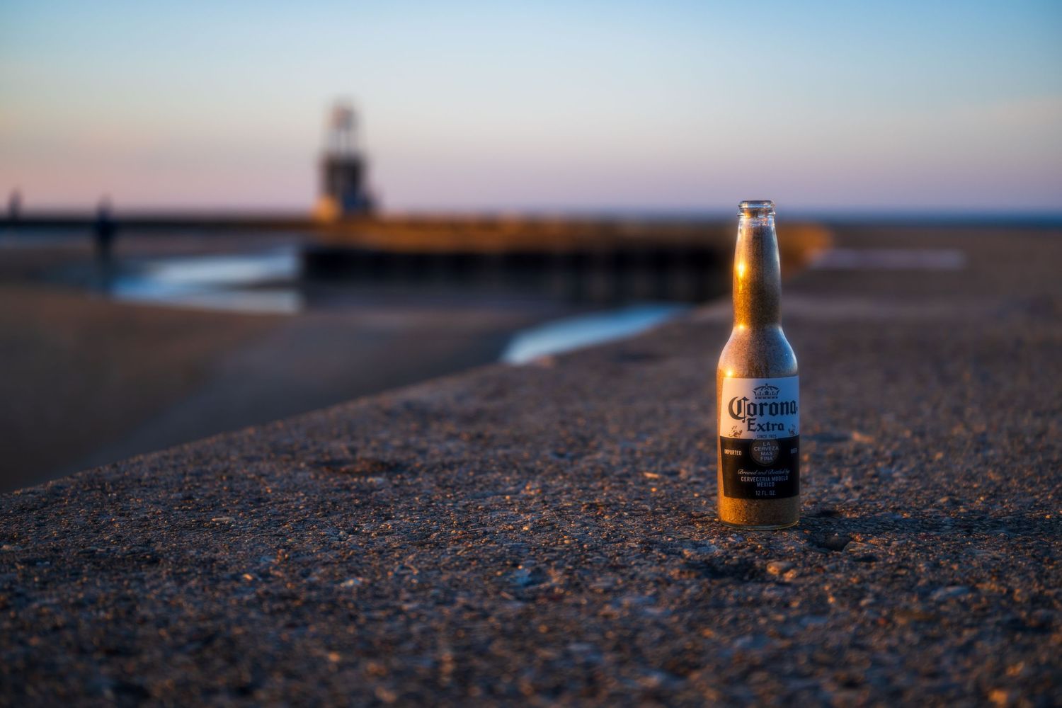 Corona Extra beer bottle on sandy beach at sunset with pier and lighthouse in background.