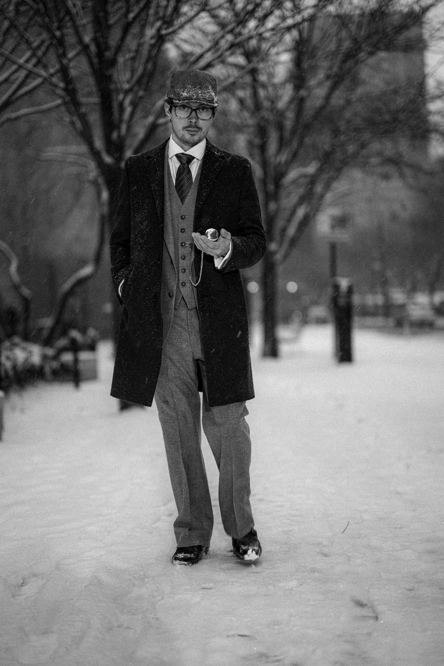 Black and white photo of a well-dressed man in overcoat and flat cap standing in snowy park.