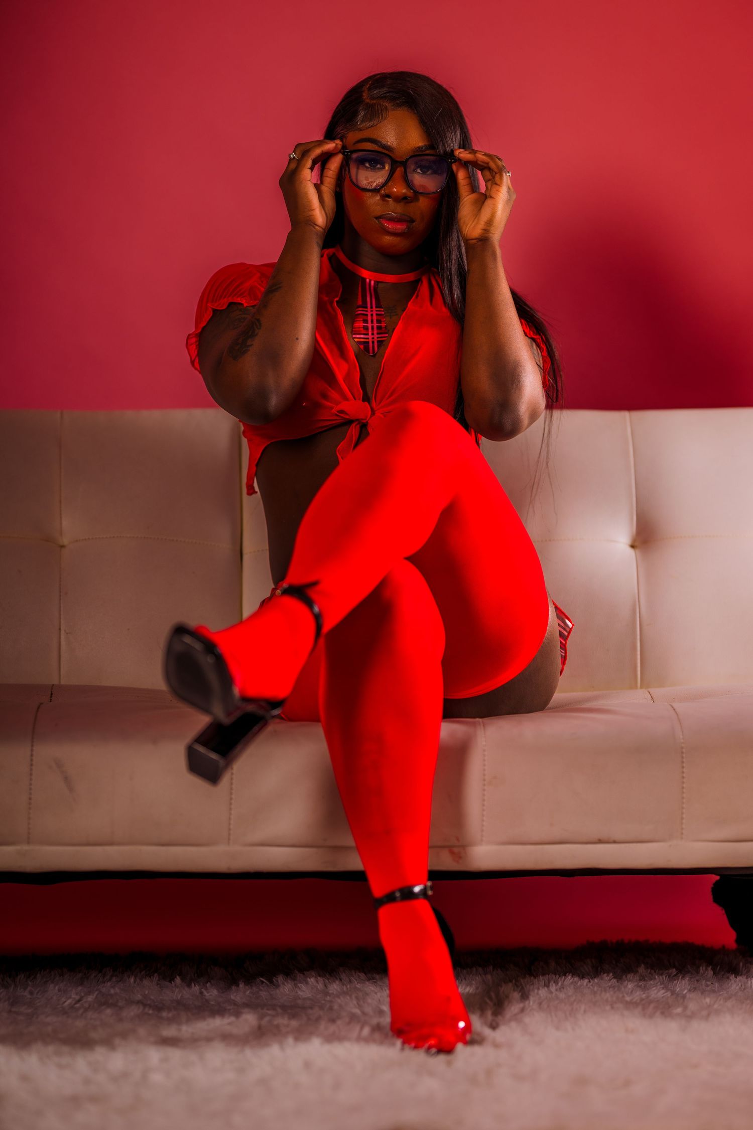 A Black woman in a red outfit with tights and heels sits on a white sofa, adjusting her glasses against a red wall.