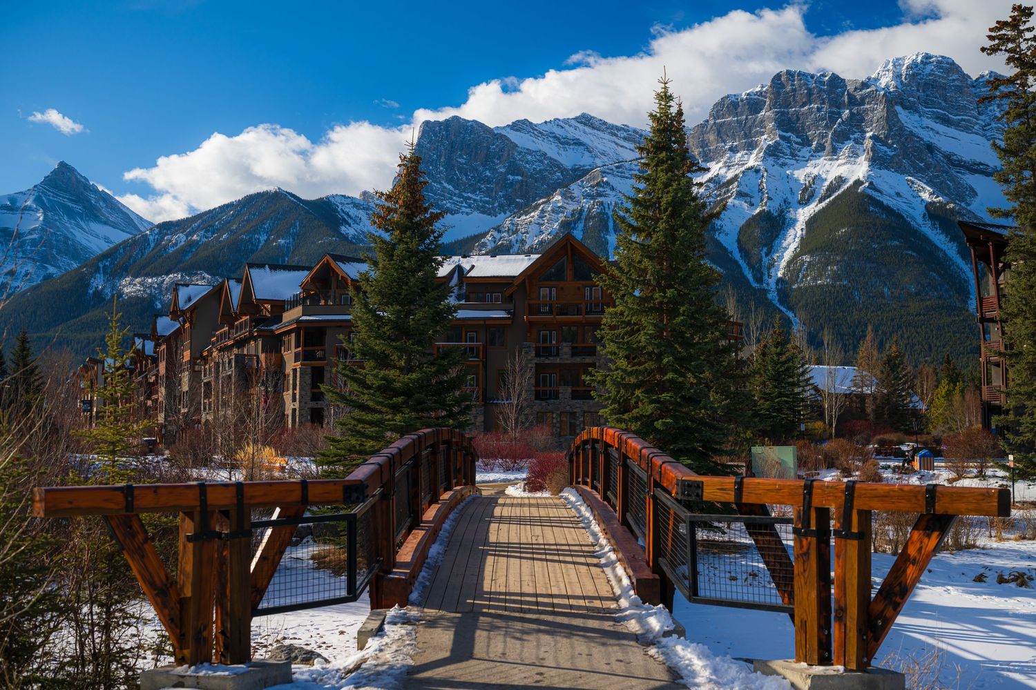Wooden bridge leading to rustic mountain lodge surrounded by snow-capped peaks and tall evergreen trees in winter.