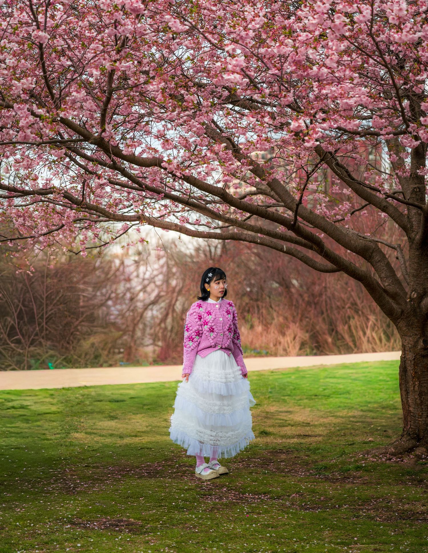 A young woman in a pink sweater and white tulle skirt stands beneath a blooming cherry blossom tree in a lush green park.