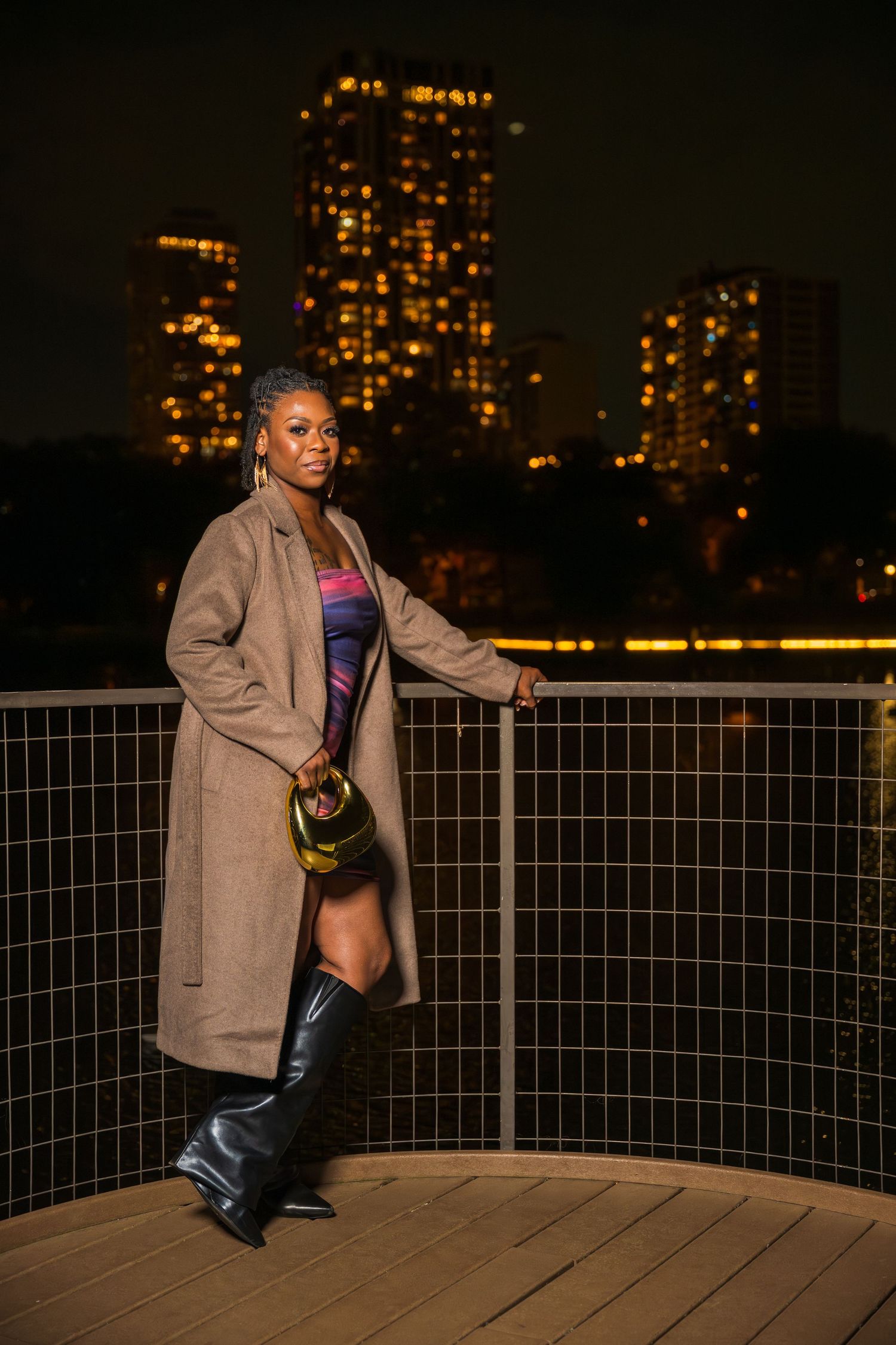 A stylish woman in a tan coat, colorful dress, and black boots poses on a city rooftop at night.