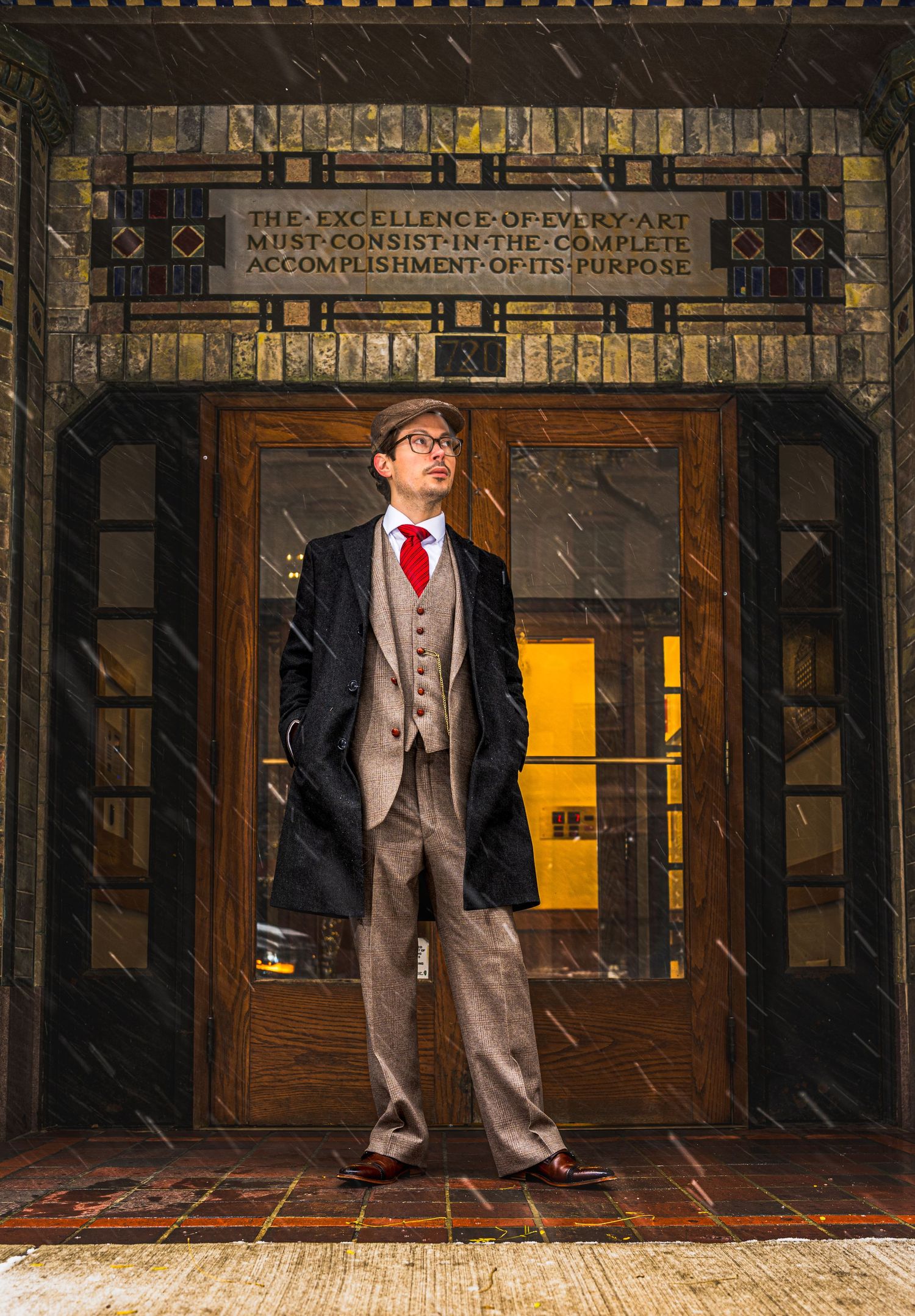 Man in vintage suit and overcoat stands before ornate building entrance with inspirational quote above doors.