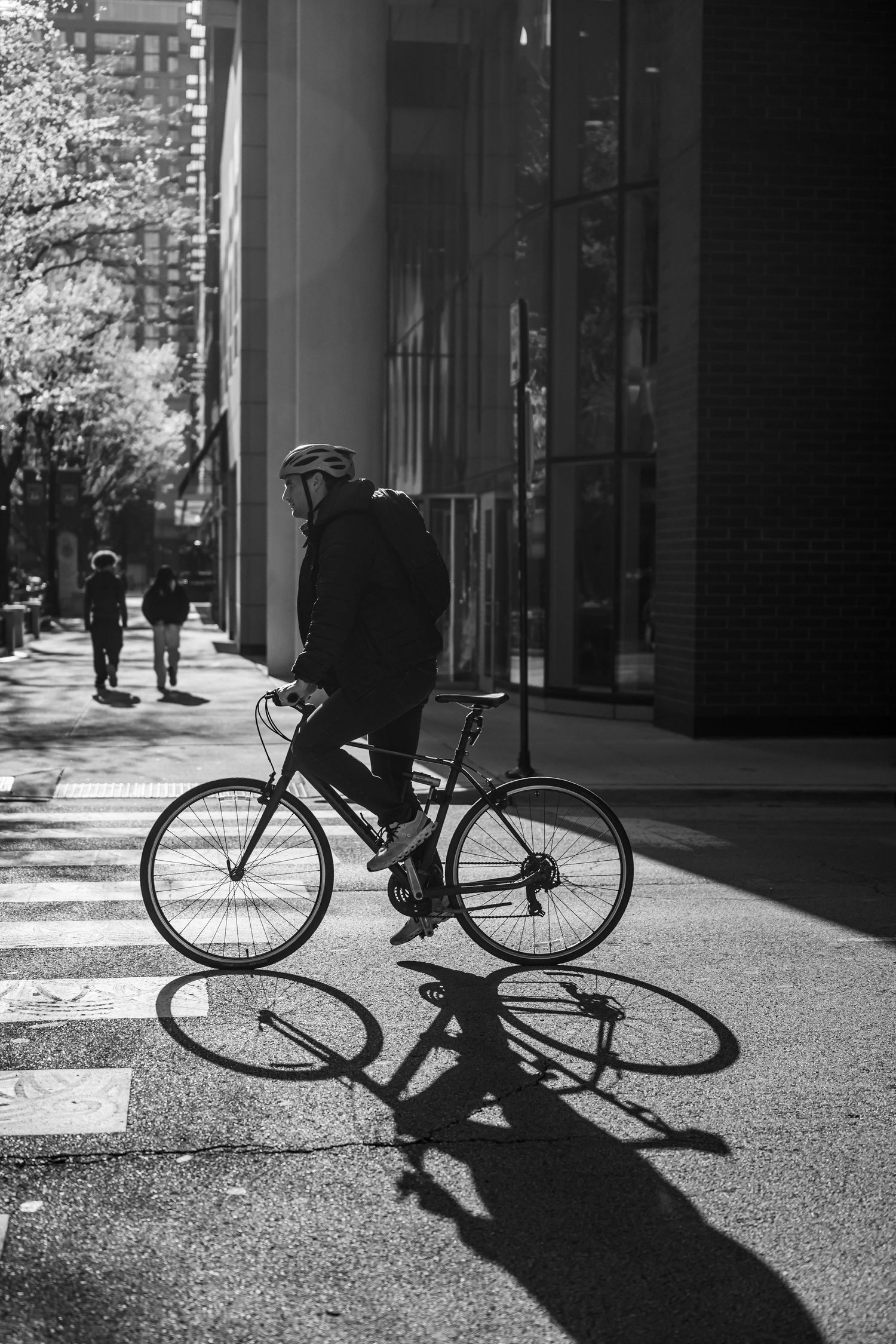 Black and white photo of a cyclist wearing a helmet pausing on a city sidewalk beside a modern building.