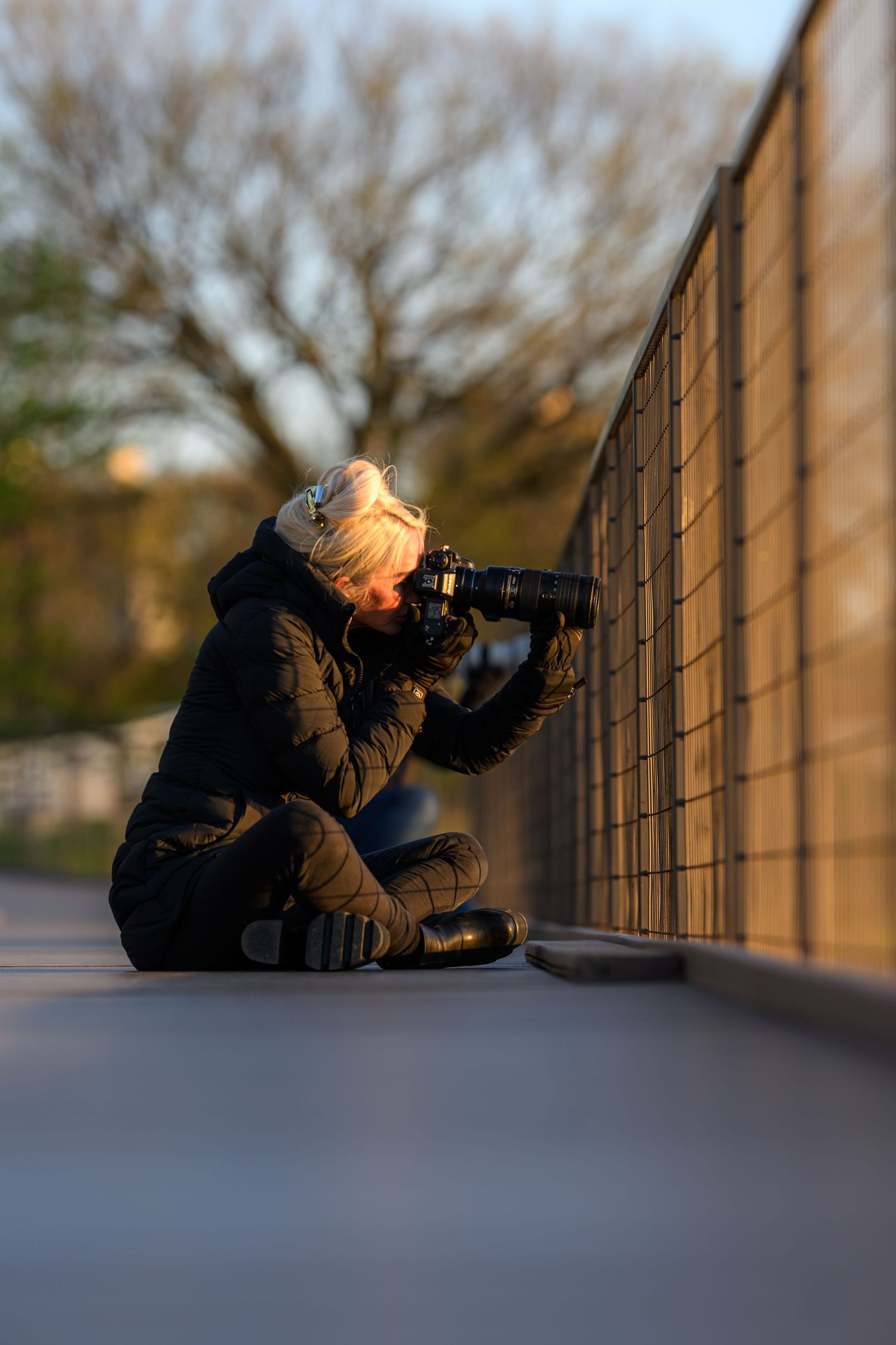 Photographer sitting on ground against fence, using DSLR camera with telephoto lens outdoors during golden hour.