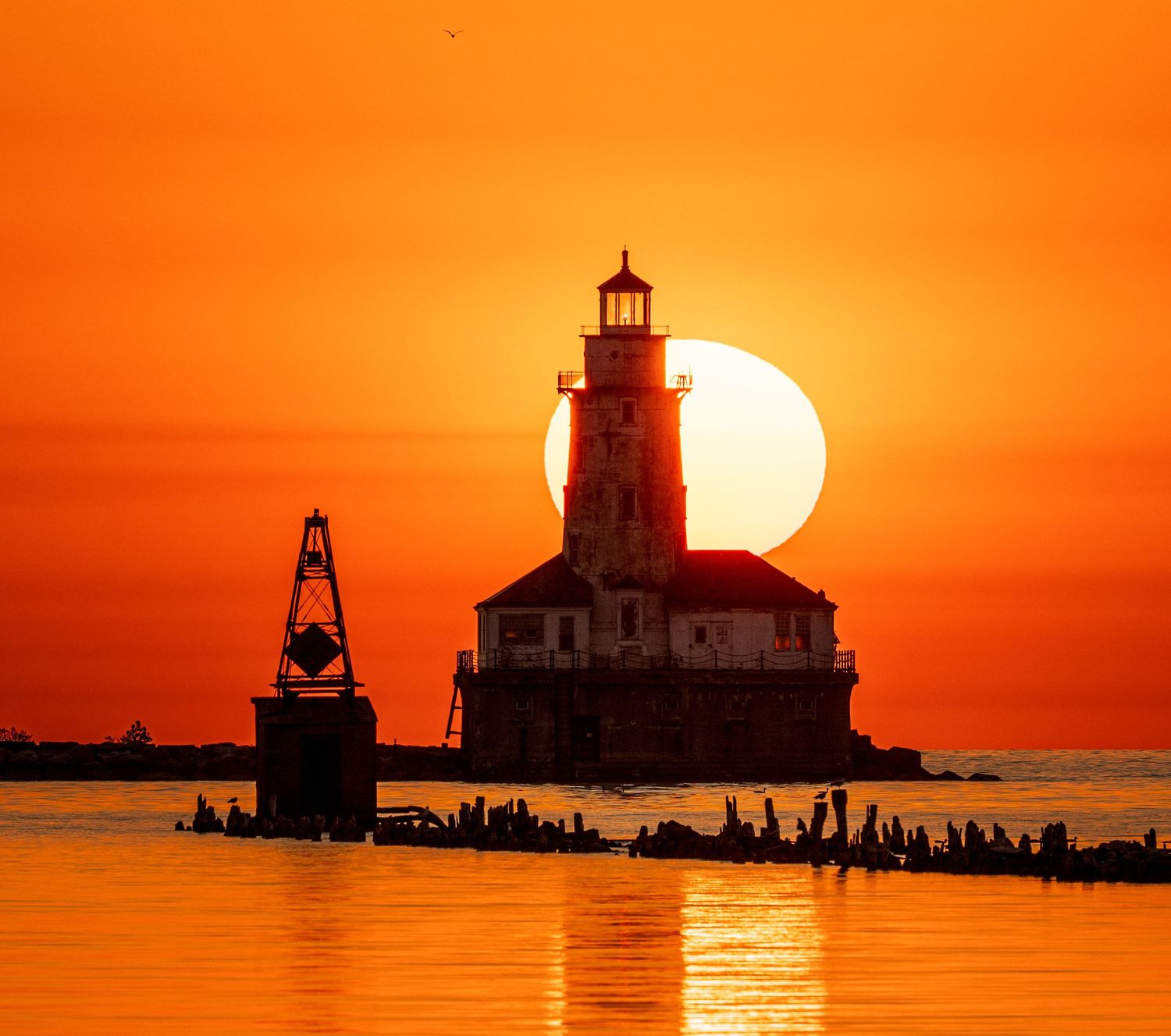 Lighthouse silhouetted against a vibrant orange sunset over calm water, with the large sun perfectly aligned behind the tower.