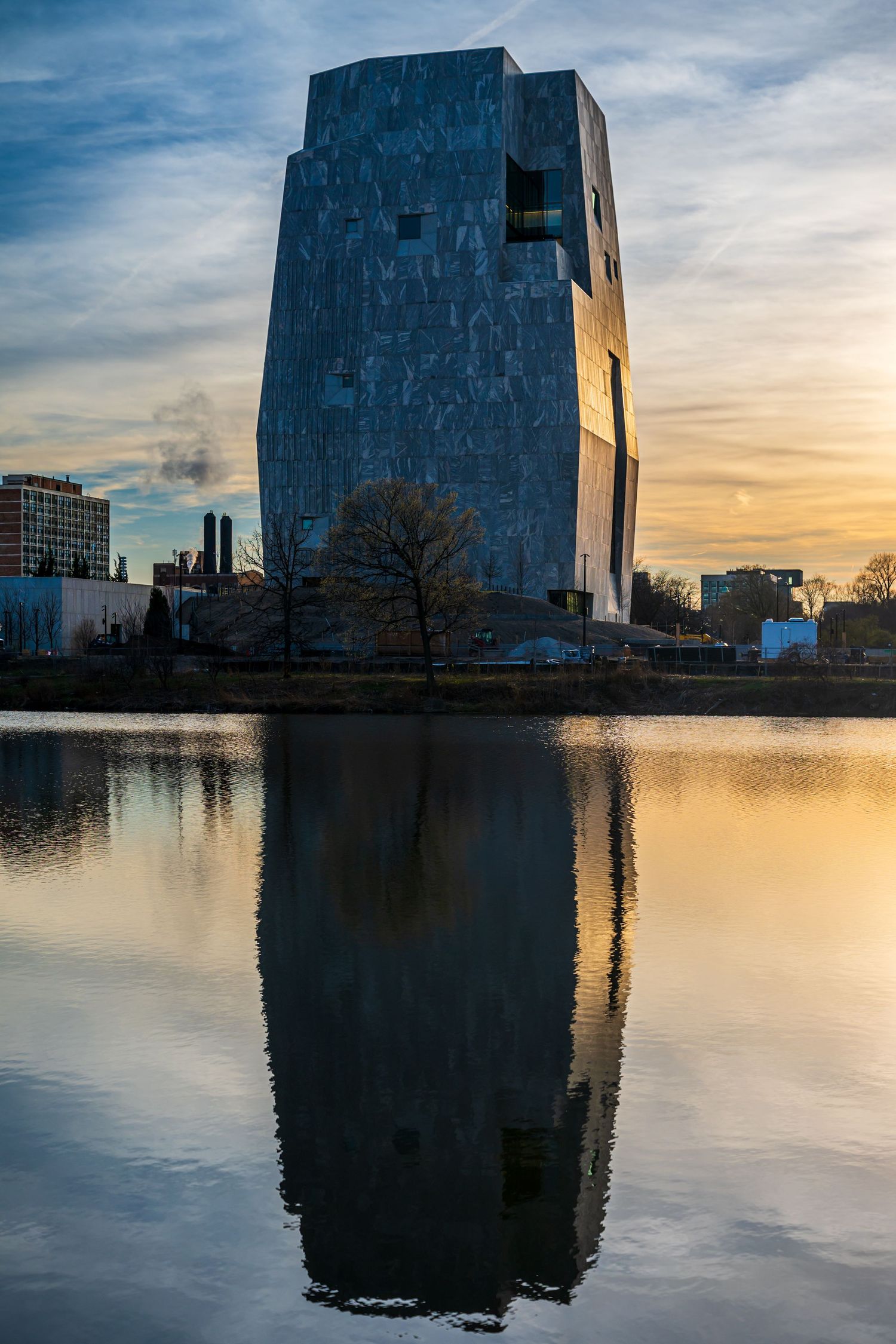 Modern blue skyscraper reflecting in calm water at sunset with dramatic clouds in Frankfurt, Germany.