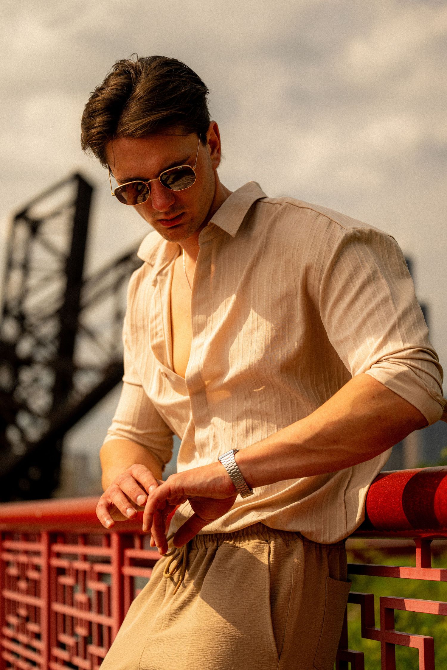 A stylish young man in a cream shirt and sunglasses checks his watch on a red bridge with an industrial backdrop.