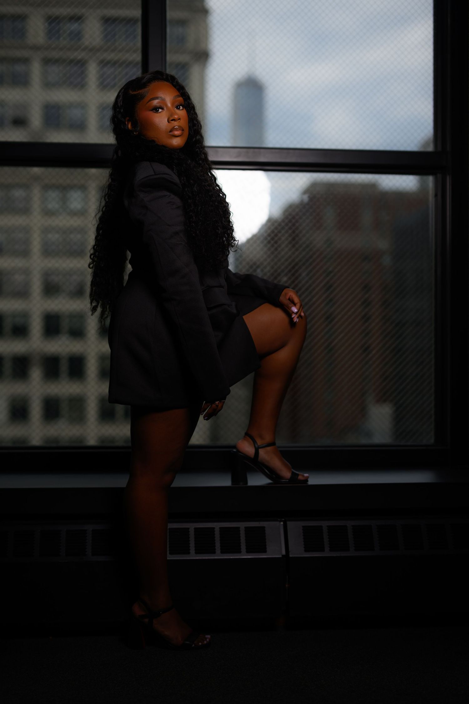 A young Black woman in a black blazer dress poses by a high-rise window with a city skyline backdrop.