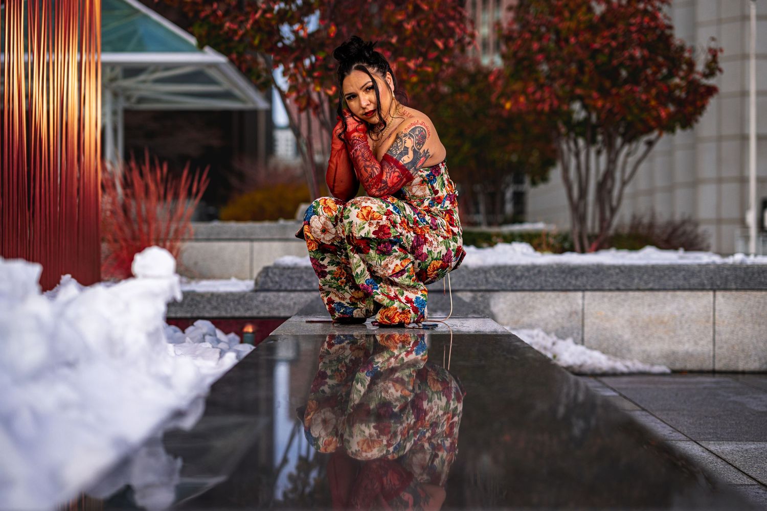 Person in colorful floral outfit crouching by reflective pool with snow and red autumn trees in background.