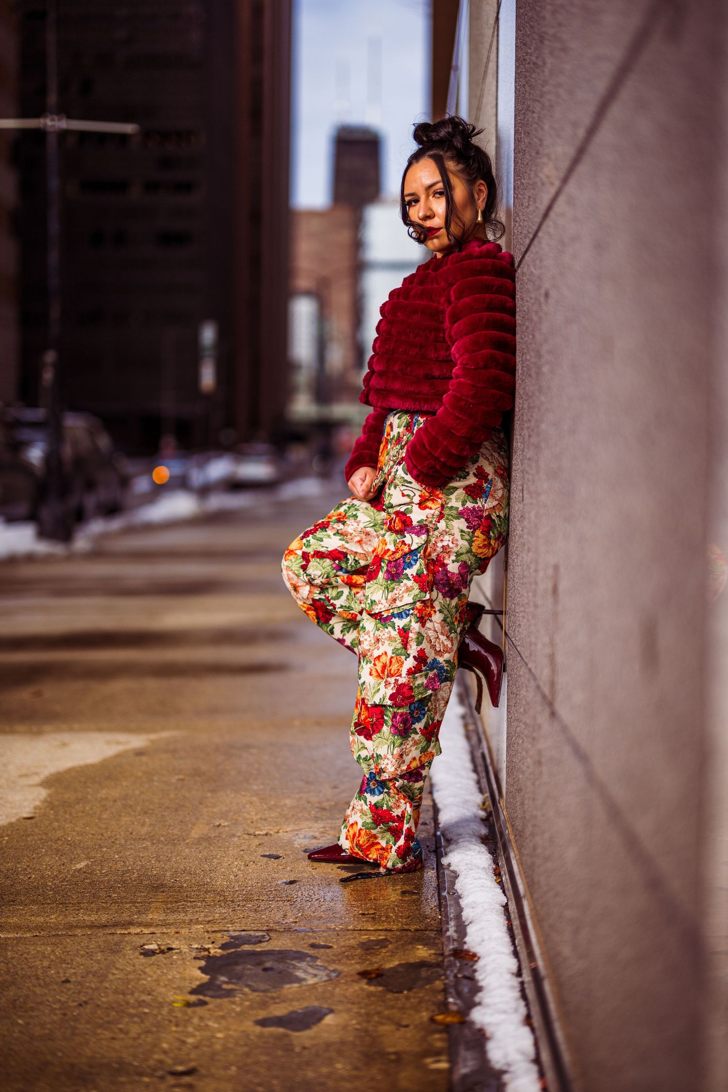 Woman in floral pants and red fur jacket leaning against urban wall on wet sidewalk with city buildings in background.