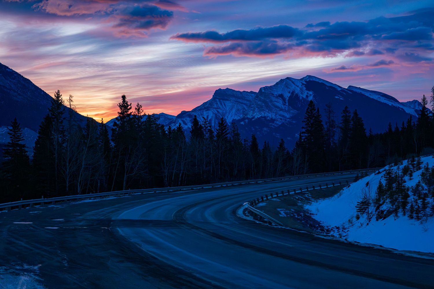 Snow-dusted mountain road curves through pine forest at twilight with vibrant purple and orange sunset sky.