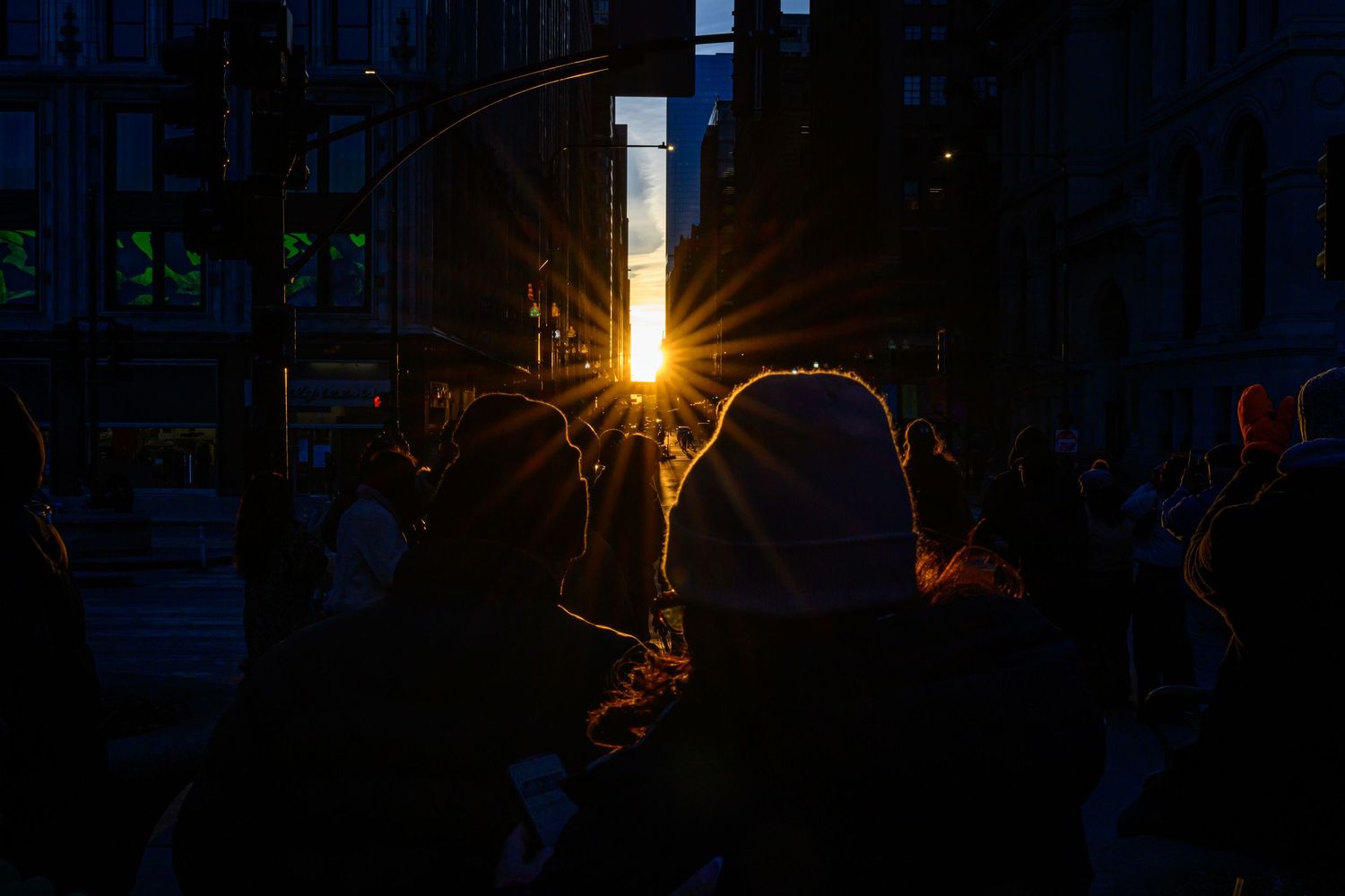 Silhouetted crowd against a stunning golden sunburst shining between dark city buildings at sunset.