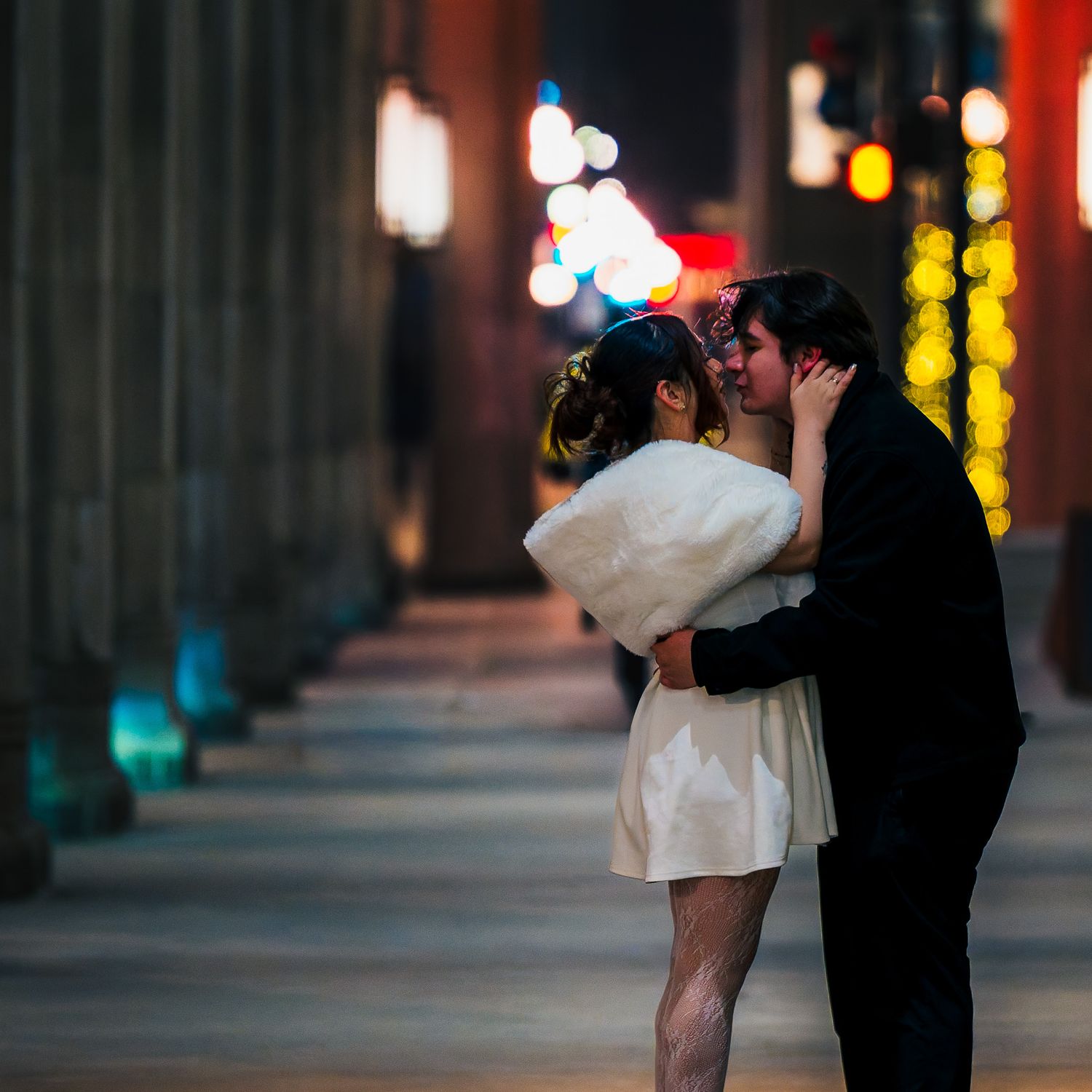 Couple sharing a romantic kiss on a city sidewalk at night, surrounded by colorful bokeh lights.